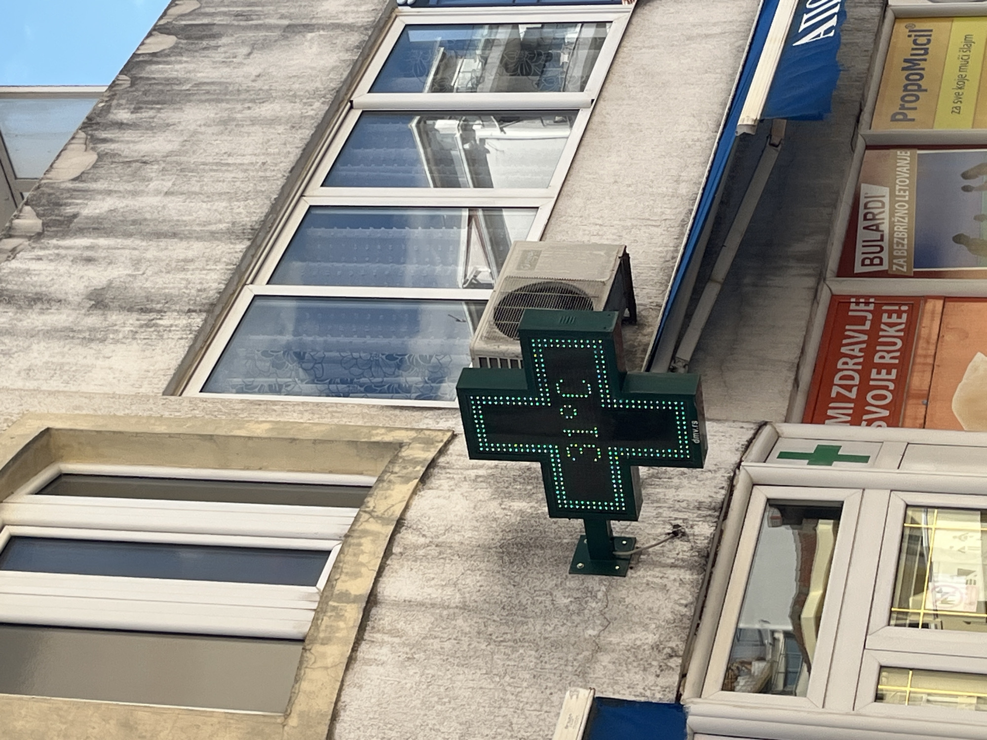 The image shows a close-up of a building facade in Budva, Montenegro, featuring a green LED cross sign displaying a temperature of 31°C. An air conditioning unit is mounted to the wall to the right of the cross. Below the cross, part of a storefront with a blue awning is visible, along with signage for "MIZDRAVLJE SVOJE RUKE!" and "BULARDI ZA BEZBRIŽNO LETOVANJE". The building's exterior is weathered concrete, and several windows with white frames are present. The sky is visible in the upper right corner.