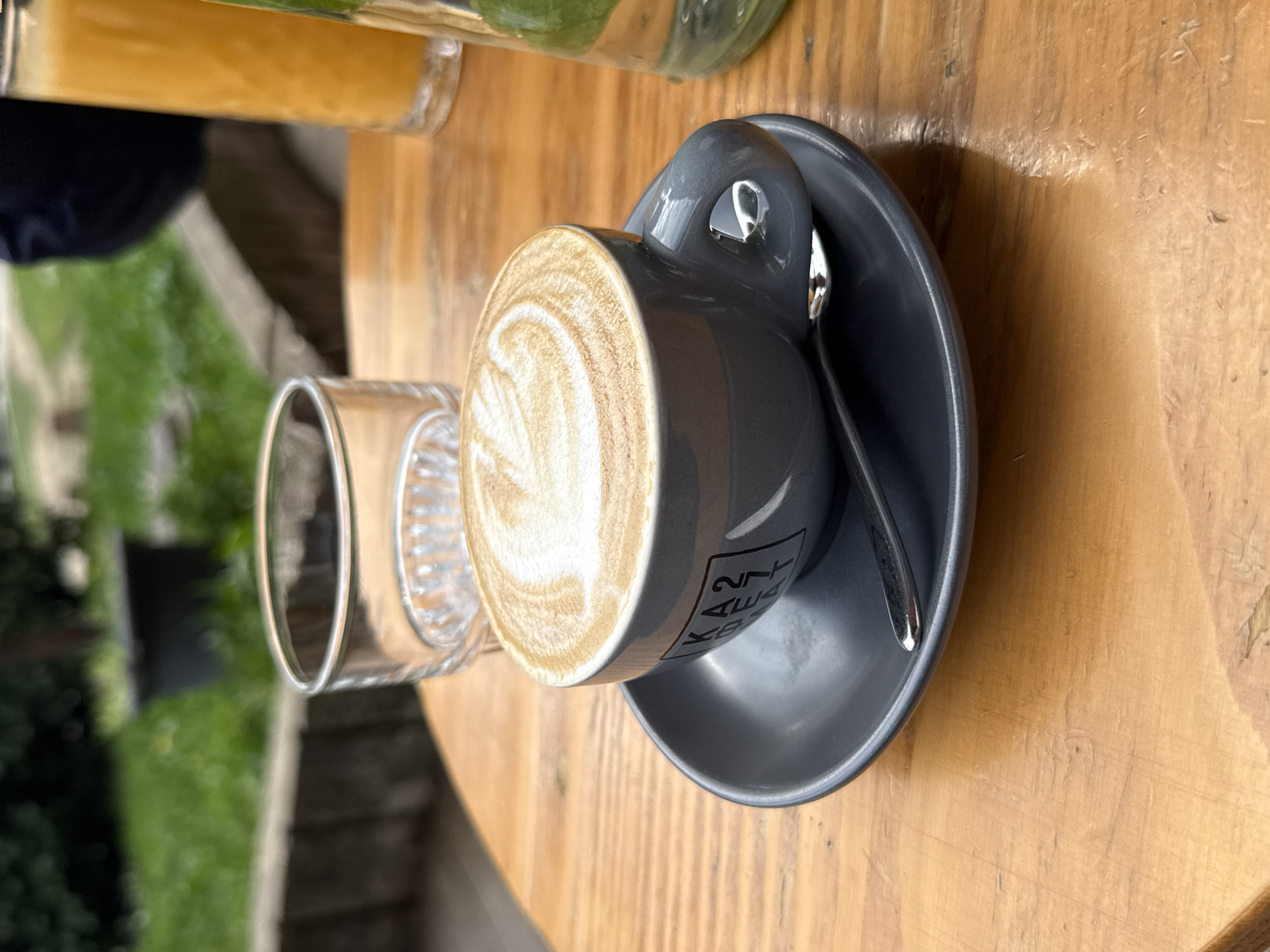 A close-up shot captures a gray coffee cup with latte art, sitting on a matching saucer with a silver spoon. Next to the cup, there's a clear glass, likely empty. The setup is placed on a wooden table, and in the background, out of focus, there is what seems to be a green bottle and outdoor greenery suggesting a cafe or restaurant setting. Based on the writing on the cup “247 kitchen and bar” this photo was taken in a cafe or restaurant in Kragujevac, Serbia.