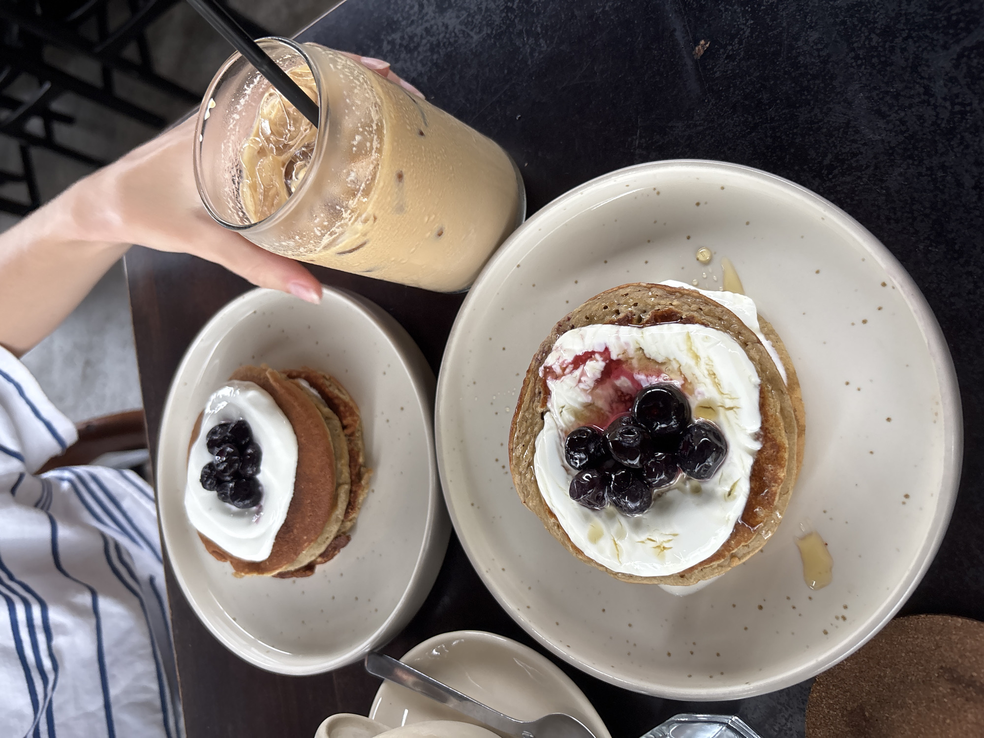 An overhead shot captures a casual meal setup on a dark table, likely in a cafe or restaurant in Kuta Selatan, Indonesia. A person's arm and hand, clad in a blue and white striped shirt, holds a clear glass of iced coffee with a black straw. The coffee is light brown and contains ice cubes.
Two plates of pancakes are prominently displayed. The larger plate, on the right, holds a stack of pancakes generously topped with white cream or yogurt, fresh blueberries, and a reddish berry jam or sauce. A golden drizzle of honey or syrup is visible on the cream and spilled onto the speckled, off-white ceramic plate. To the left, a smaller plate features a stack of pancakes also topped with white cream and a cluster of blueberries. Both pancake servings appear to be a light brown color.
A small, empty ceramic dish with a silver spoon rests near the larger pancake plate. A circular, light brown cork coaster is partially visible at the bottom right. The scene suggests a relaxed breakfast or brunch setting, with the food prepared for consumption. No specific time of day or weather is discernible from the indoor setting, and no visible text is present.
