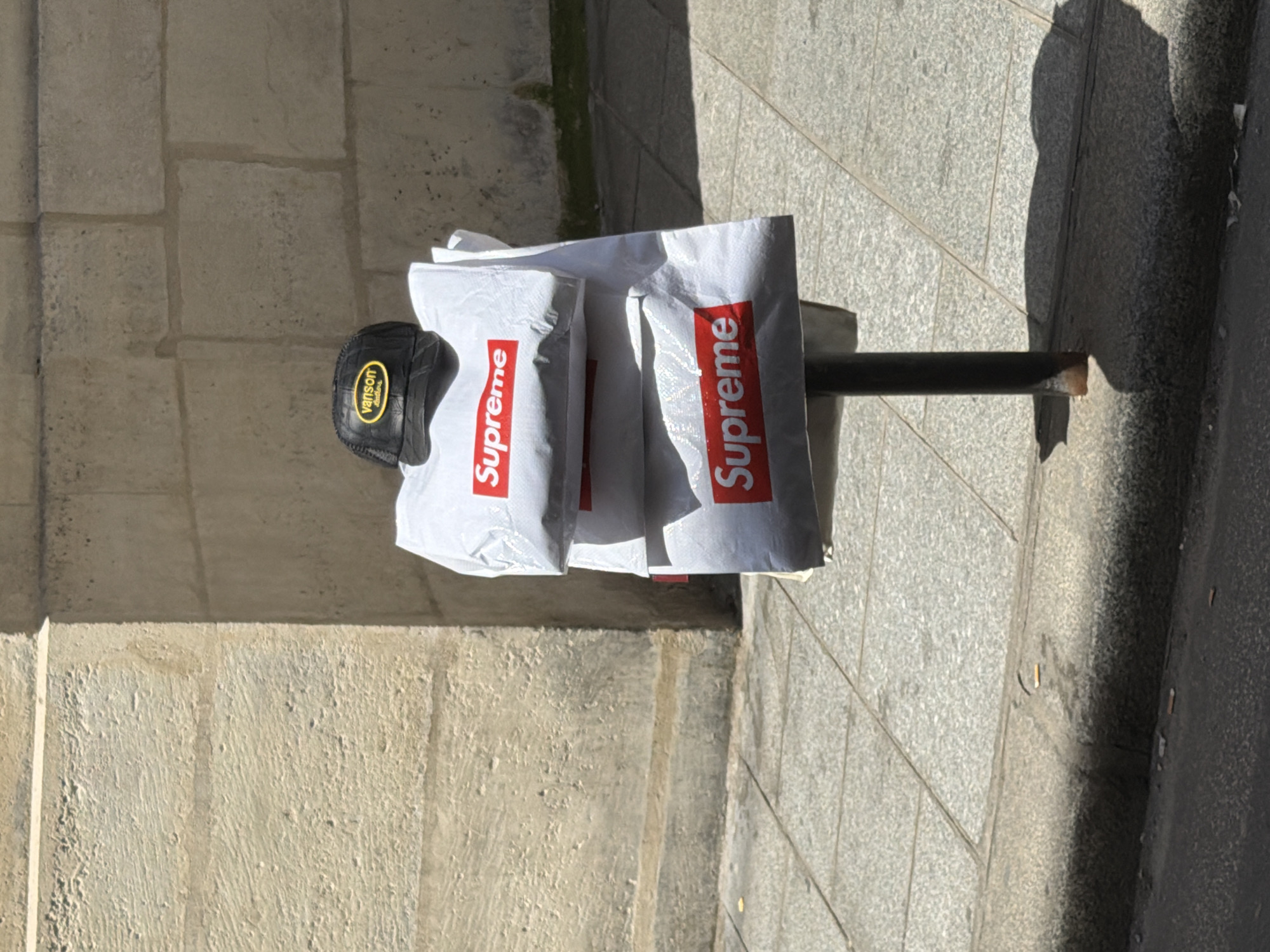 The image shows a hat and three white shopping bags with red "Supreme" logos resting on a metal bollard or post. The hat, which appears to be black with a mesh back and a yellow "Vanson Leathers" logo, is positioned to the left of the bags. The bollard is matte dark grey, and is located on a paved sidewalk or pedestrian area next to a stone wall. The image was taken outside in a well-lit, sunny setting. Based on the type of stonework in the wall and the bollard, the image may have been taken in Paris, France.