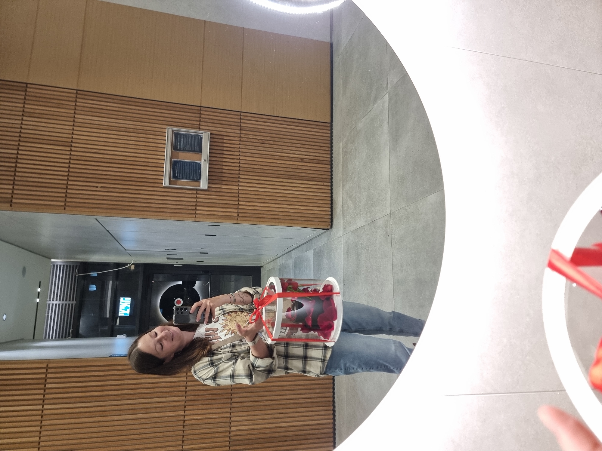 A woman is standing in what appears to be an upscale interior space, possibly a building in Tel-Aviv, Israel. She is holding a clear, cylindrical container filled with red roses, secured with a red ribbon. The woman is wearing a plaid shirt, a graphic tee, and jeans. She is holding a smartphone, presumably taking a photo of herself with the roses.
The environment consists of a combination of materials: wood panels on the wall, smooth, possibly tiled floors, and the white, curved shape of an architectural element overhead, possibly a part of a sculpture or building design. There is also a hallway with illuminated signage visible in the background.