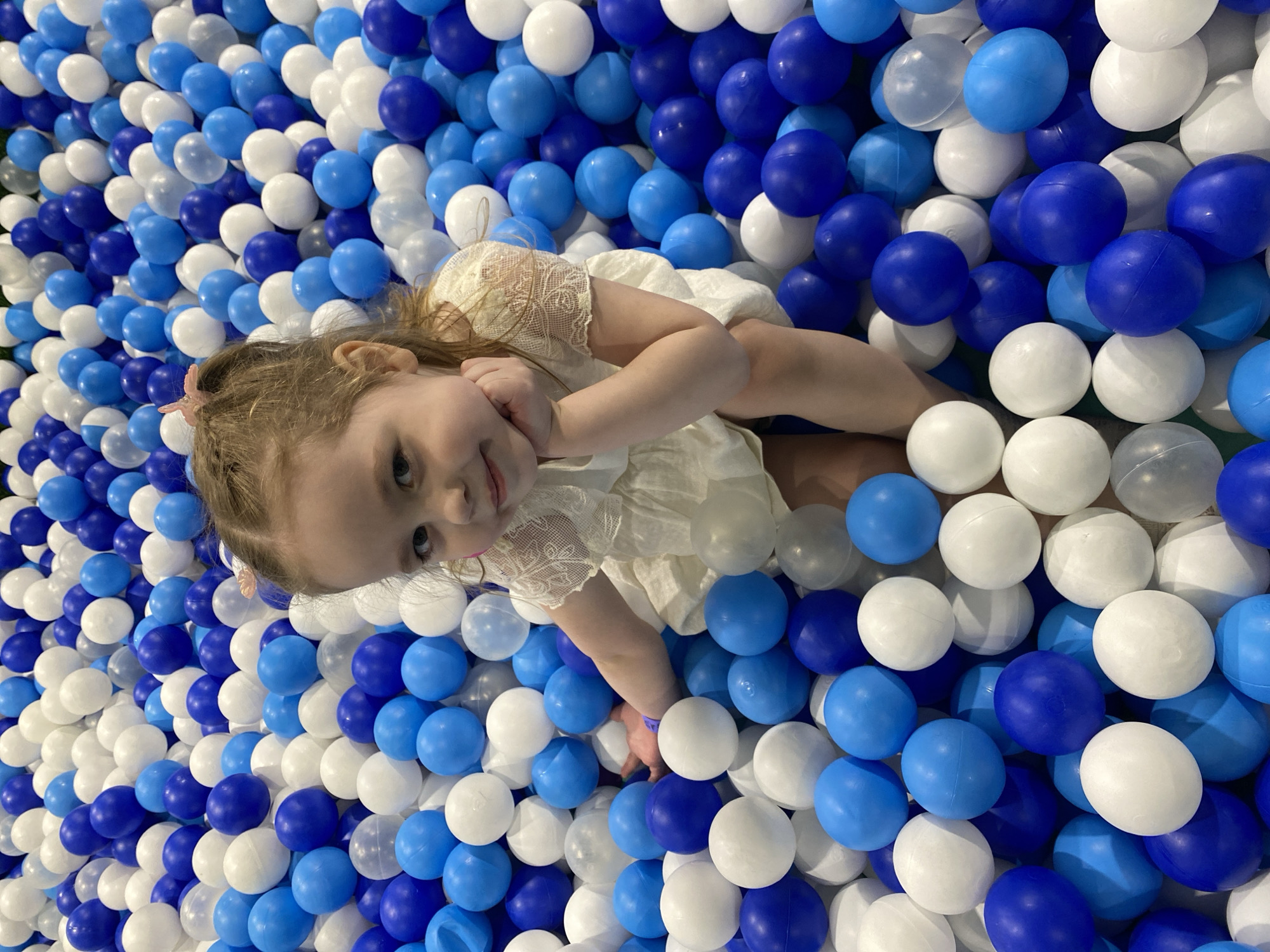 This image captures a young child, likely a girl, lying on her back within a vibrant ball pit. The child has light-colored hair, which features a small pink hair clip on her left side, and she is dressed in a light-colored (possibly white or cream) dress with delicate lace detailing on the shoulders. She gazes upwards towards the camera with a gentle, content expression, a soft smile playing on her lips. Her right arm is bent with her hand resting near her face, while her left arm is also bent and immersed among the balls.
The scene is dominated by a large quantity of plastic balls in various shades of blue—ranging from light to dark—interspersed with white balls. These balls completely fill the frame around the child, indicating an indoor ball pit, common in children's play areas or entertainment centers. The visual texture created by the myriad of spheres is prominent. No specific time of day is evident, but the setting is clearly indoors. There is no visible text in the image. The overall impression is one of leisure, enjoyment, and comfort in a playful environment located in Izhevsk, Russia.