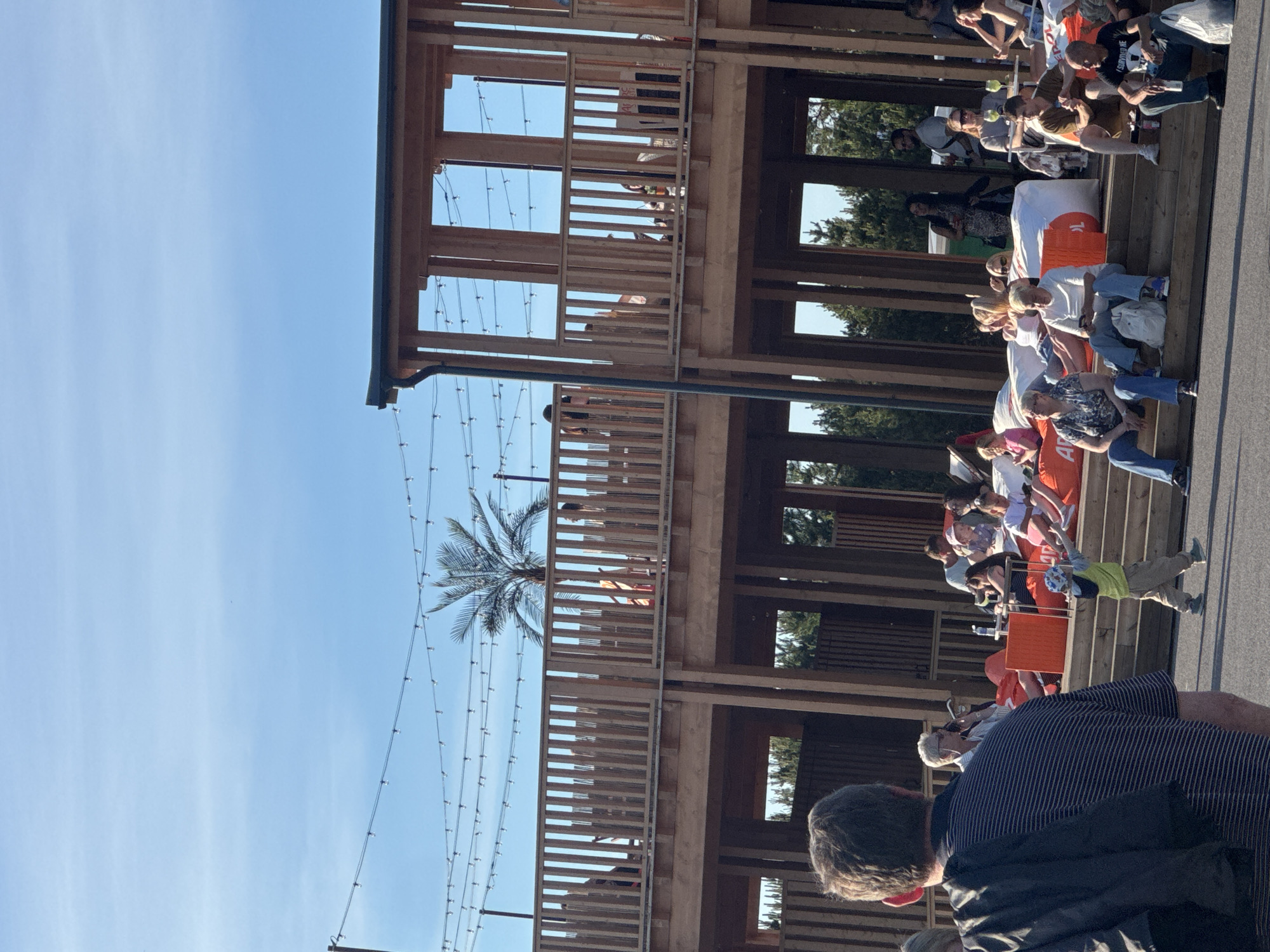 The image depicts an outdoor seating area with people relaxing on large orange beanbag chairs arranged on tiered wooden steps. Several individuals are seated, some are talking, and others appear to be observing their surroundings. The scene is set against a backdrop of a modern, multi-level wooden structure with open-air balconies and railings. String lights are visible overhead, suggesting an evening or late afternoon setting. A palm tree is present in the background, along with other greenery. The sky is clear and blue. In the foreground, a person wearing a striped shirt is partially visible, and a young child in a yellow shirt and a blue hat is walking on the asphalt in front of the seating area. This appears to be a public recreational or dining space, potentially part of a complex with multiple levels, in the city of Tallinn, Estonia.