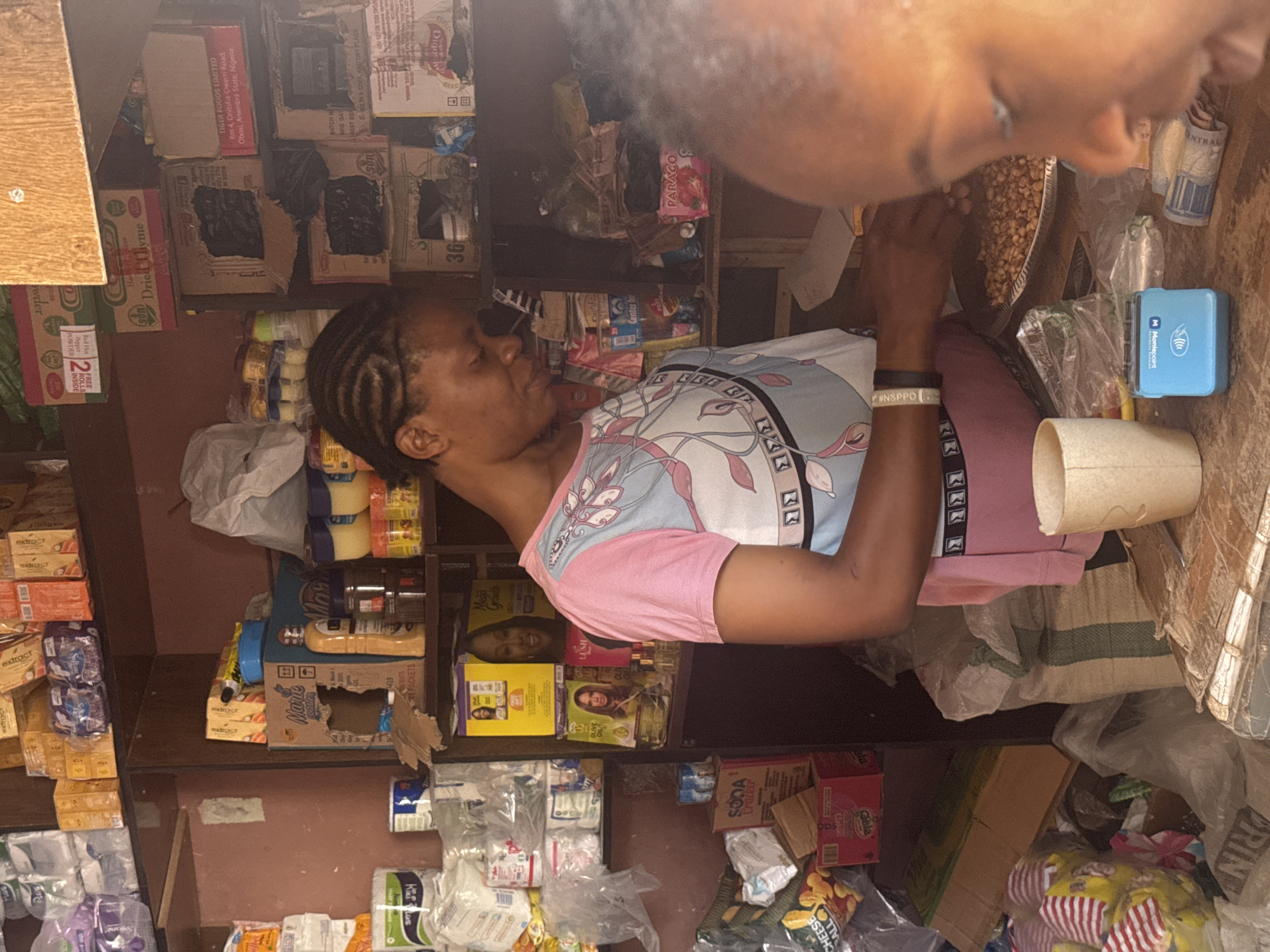 The image depicts an indoor scene, possibly a small shop or market stall, with shelves filled with various products. In the foreground, a woman with braided hair, wearing a pink and blue patterned shirt and pink leggings, is engaged in an activity that involves handling small, light brown items from a silver platter. Her hands are focused on this task. To her right, a portion of another person's face, likely a child, is visible in close proximity, looking towards the center of the scene. On the wooden counter in front of the woman, there is a light blue point-of-sale device and some folded currency. The environment appears to be somewhat cluttered, with various packaged goods, bags, and boxes on shelves and the floor. The lighting suggests it might be daytime, with natural light contributing to the scene. The overall impression is of a transactional environment where goods are being prepared or handled. Based on the presence of the "Tiger Foods Limited" box indicating "Obosi, Anambra State, Nigeria" as a manufacturer, this scene likely takes place in a commercial setting within Nigeria, potentially in a city like Abuja, given the nature of small retail businesses in such locations.