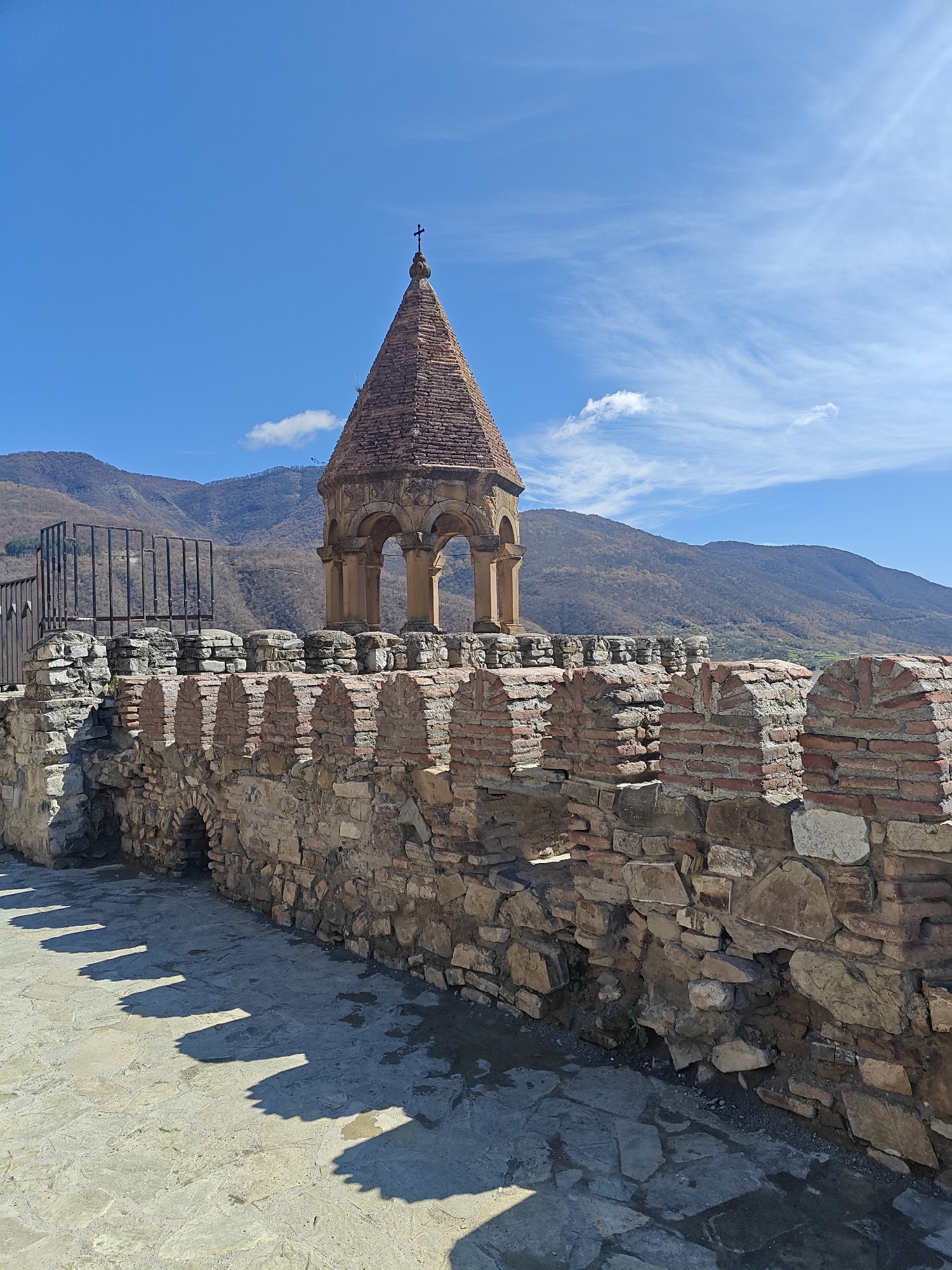 The image shows a stone and brick fortification wall topped with a tower. The tower is capped with a cross. In the background are mountains under a blue sky with scattered clouds. The location is an unknown city in an unknown country.