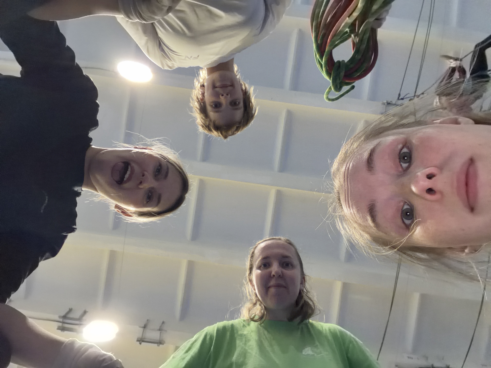 Five individuals are positioned in a circle, their faces directed downwards toward the camera, creating a "worm's-eye view" perspective. The setting appears to be indoors, identifiable as a climbing gym in Berezniki, Russia based on circumstantial information. The background is a white ceiling with fluorescent lights and mounted climbing hardware. From the bottom left moving clockwise, the first person is wearing a black shirt and their mouth is open. The second person is wearing a green shirt. The third person has blond hair. The fourth person is holding some green and red rope and wearing a white shirt. The fifth person is cropped out of the image, only the edge of their face is visible.