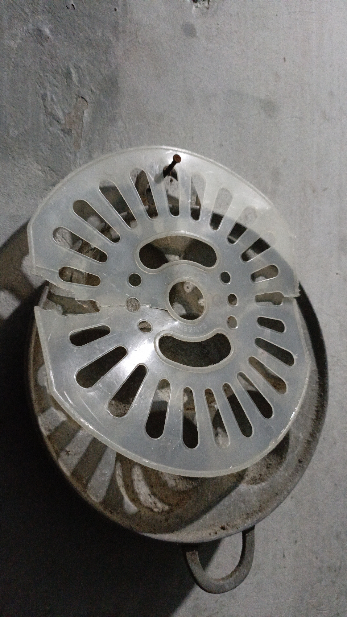 A rusty metal pot with a broken white plastic strainer on top sits on a gray concrete surface. A small nail protrudes from the plastic strainer. The background is a gray, textured wall. The scene appears to be outdoors in Trihanggo, Indonesia, possibly in a residential area or a workshop.