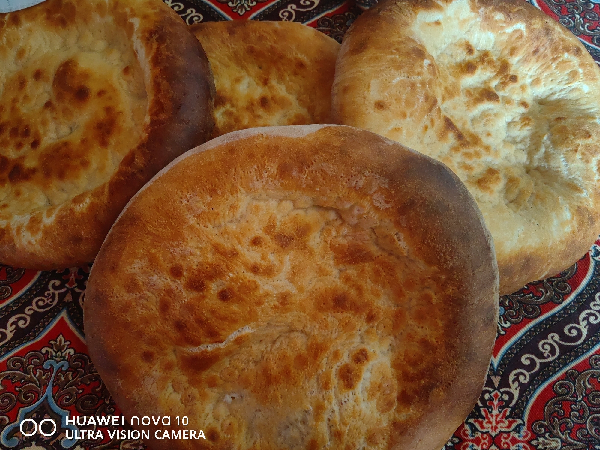 A close-up, overhead shot displays four flatbreads of varying sizes and shades of golden brown, arranged on a patterned fabric. The fabric has a red base with intricate black, white, and blue swirling designs. One of the flatbreads in the foreground is larger and has a slightly darker, more browned edge. The other three are positioned behind and to the sides, with textures ranging from smooth to slightly bumpy. A watermark at the bottom left corner reads "HUAWEI NOVA 10 ULTRA VISION CAMERA." The scene is indoors, likely in a domestic setting, and the lighting suggests a natural light source, possibly from a window. There are no people or animals visible in the image. The location is identified as Jetim-Dobo, Kyrgyzstan, implying this is a typical display of bread from that region.