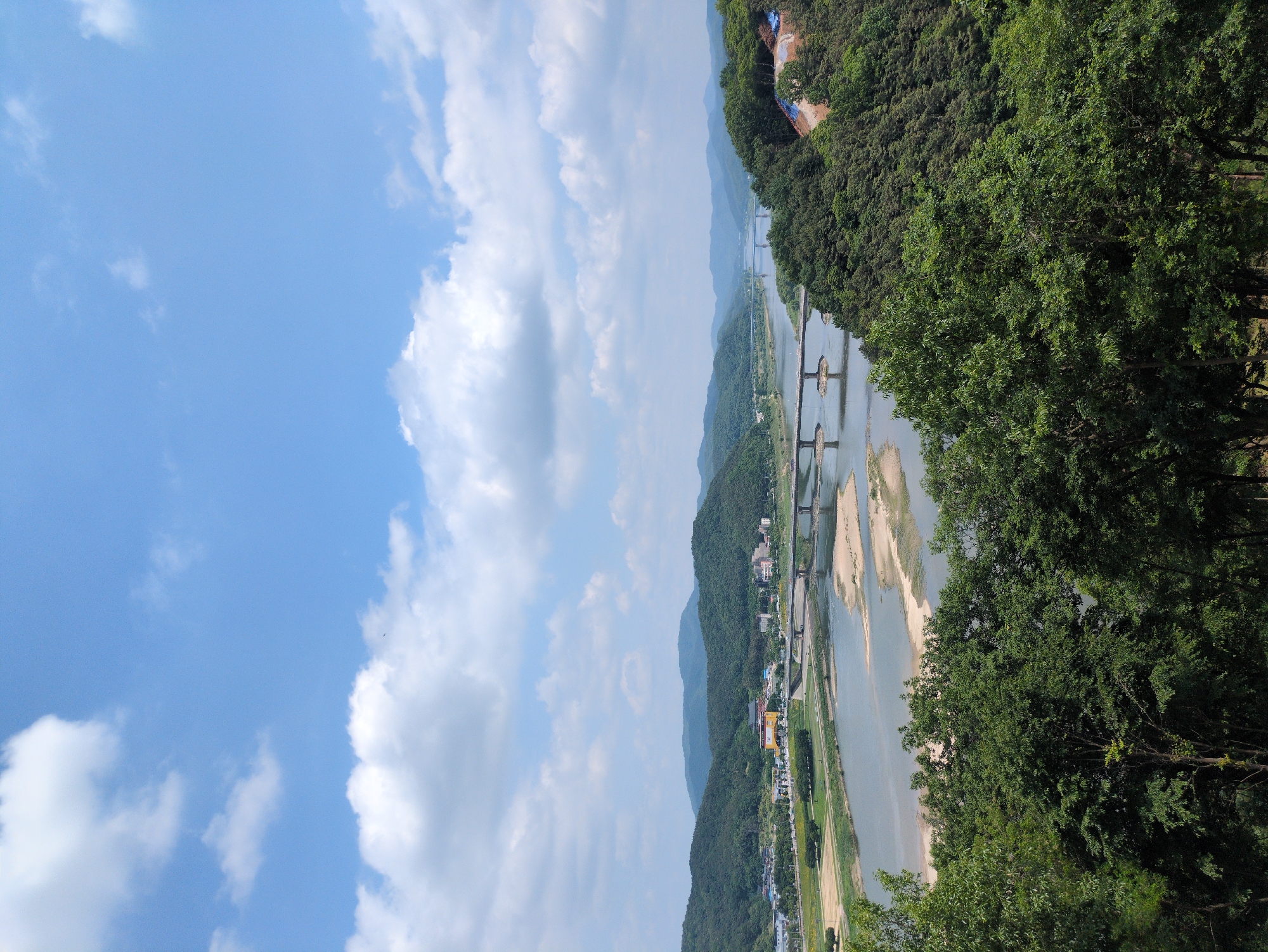 The image shows a landscape view in Gongju-si, South Korea. The scene is dominated by a wide river with sandbanks and a series of bridges spanning it. Lush green mountains form the backdrop, with a section of a town visible along the riverbank. The sky is blue with scattered white clouds. Trees are present in the foreground, partially obscuring the view.