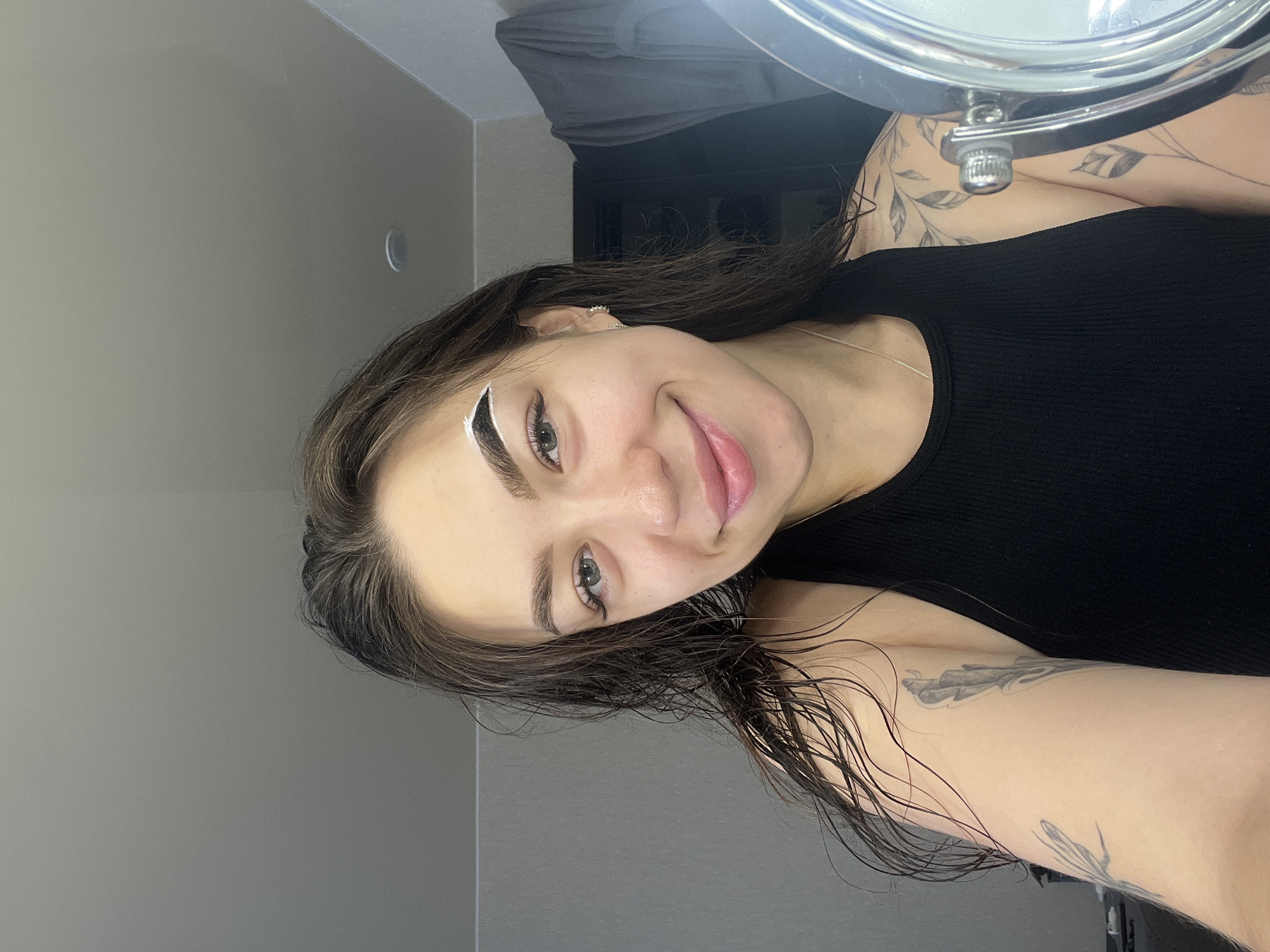 In a dimly lit room, a young woman with wet dark hair is taking a selfie in a mirror. She is wearing a black tank top and has tattoos on her arms. Her left eyebrow has white product applied to it, suggesting she might be dyeing or shaping her eyebrows. The background shows a gray wall, a closet door, and a towel hanging on a rack. The lighting is soft and appears to be from artificial sources. The scene does not contain any information that places it specifically in Chaikovsky, Russia.