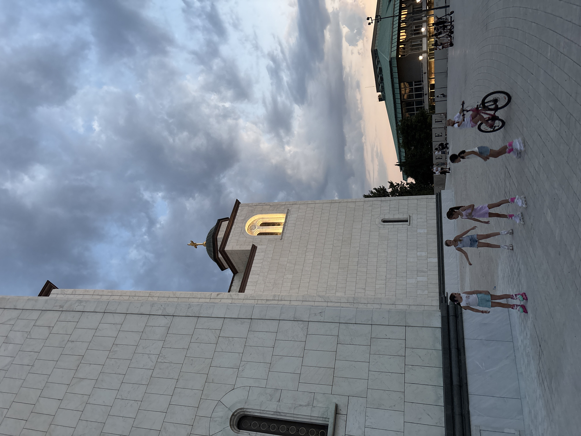 The image depicts an outdoor scene in Belgrade, Serbia, likely in front of the Church of Saint Sava. In the foreground, four young girls are rollerblading on a paved plaza. The girl on the far left is wearing a white tank top and blue shorts. The second girl from the left is wearing a white tank top and denim shorts. The third girl from the left is wearing a light pink dress. The girl on the far right is riding a pink bicycle. Behind the girls, a large, modern building with a green roof is visible. The sky is filled with dramatic, cloudy formations, suggesting twilight or a storm. The architecture of the Church of Saint Sava is prominent on the left side of the image, characterized by its white marble facade and a distinct dome topped with a cross.