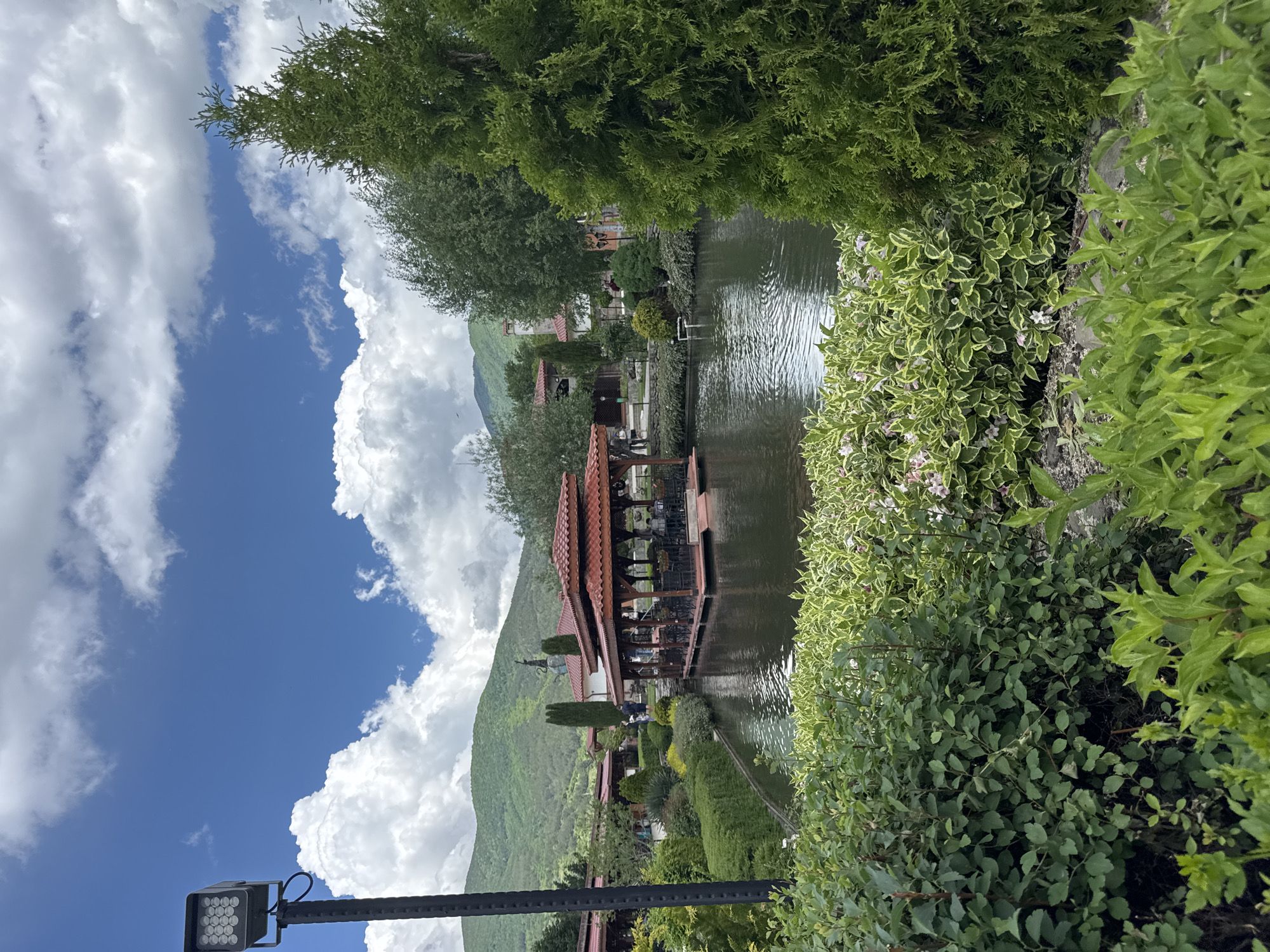 The image depicts a tranquil scene in the city of Skobelevo, Bulgaria. A structure with a red-tiled roof is situated in the middle of a body of water, possibly a lake or a large pond. The structure appears to be a gazebo or a small restaurant. Around the water, there are various types of greenery, including trees, bushes, and flowering plants. A mountain rises in the background, partially obscured by large, puffy clouds in a blue sky. A street light is visible in the foreground. There are people present in the structure on the water.