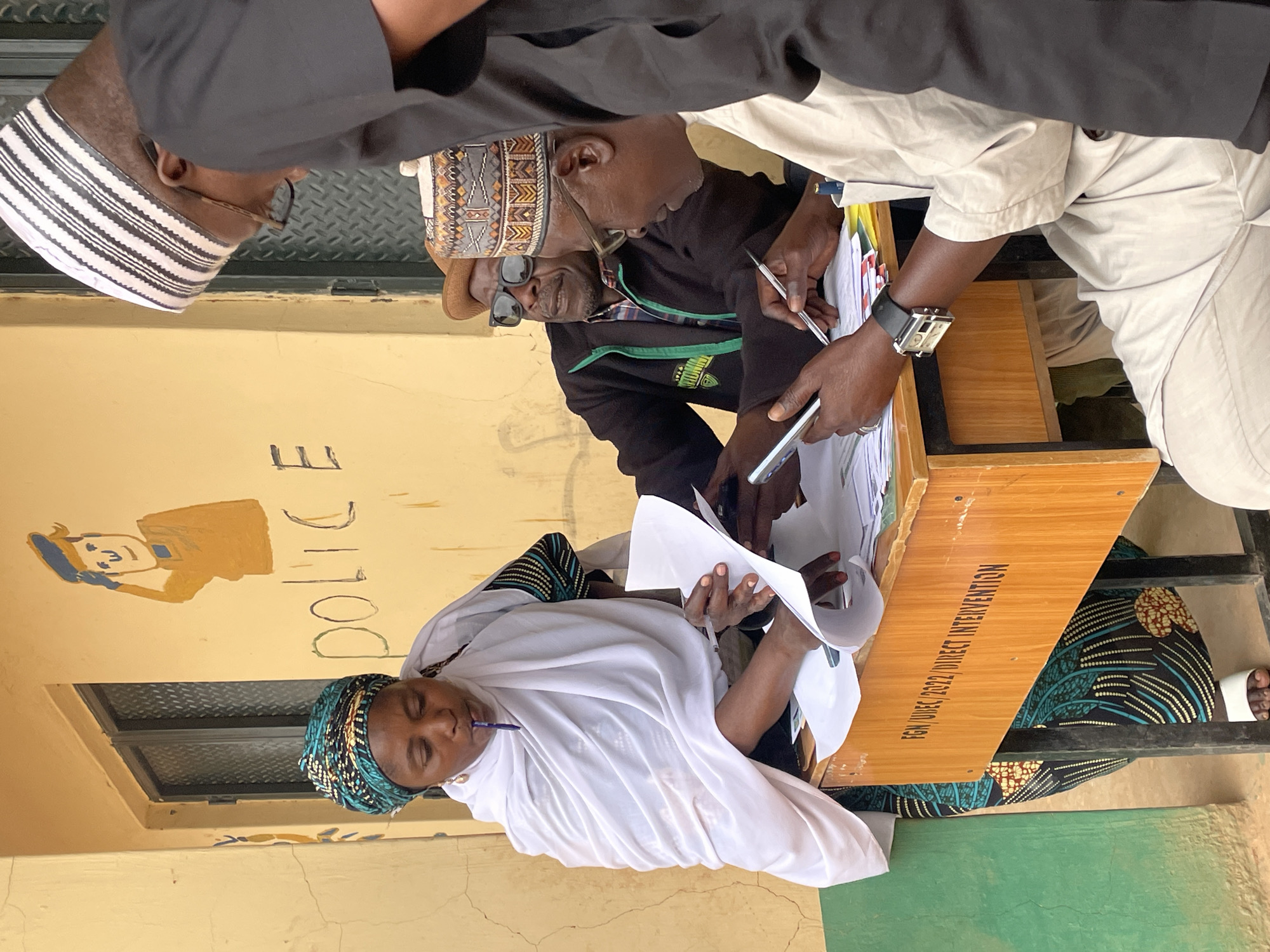 Here is a detailed description of the image:
**Scene**: An indoor scene in Jos, Nigeria, shows a group of people gathered around a desk. A woman in a white garment and patterned headscarf sits at the desk, holding papers and a pen in her mouth. Three men are standing or leaning over the desk, seemingly observing or assisting the woman.
**People**:
* Woman: Wearing a white garment, patterned headscarf, holding papers, and a pen.
* Man 1: Wearing a black cap with white stripes, glasses.
* Man 2: Wearing a decorated brown cap, sunglasses, a black jacket, and a green shirt.
* Man 3: Wearing a khaki colored top, holding a pen and phone, and sporting a watch.
**Objects**:
* Desk: Made of wood. The desk's surface has text "FGN/UBEC/2022/DIRECT INTERVENTION" written in black.
* Papers: Numerous sheets held by the woman and lying on the desk.
* Pens: Held by the woman and one of the men.
* Watch: Visible on one of the men's wrists.
* Phone: Being held by one of the men.
* Wall: Painted yellowish with an illustration of a police officer and the word "POLICE".
**Actions/Interactions**: The woman appears to be filling out or reviewing documents, with the men observing and possibly assisting her. The men are holding pens, indicating that they may be involved in the process.
**Environment**: The scene takes place indoors. The background includes a window above the woman and what appears to be a wall painted with an illustration of a police officer and the word "POLICE".