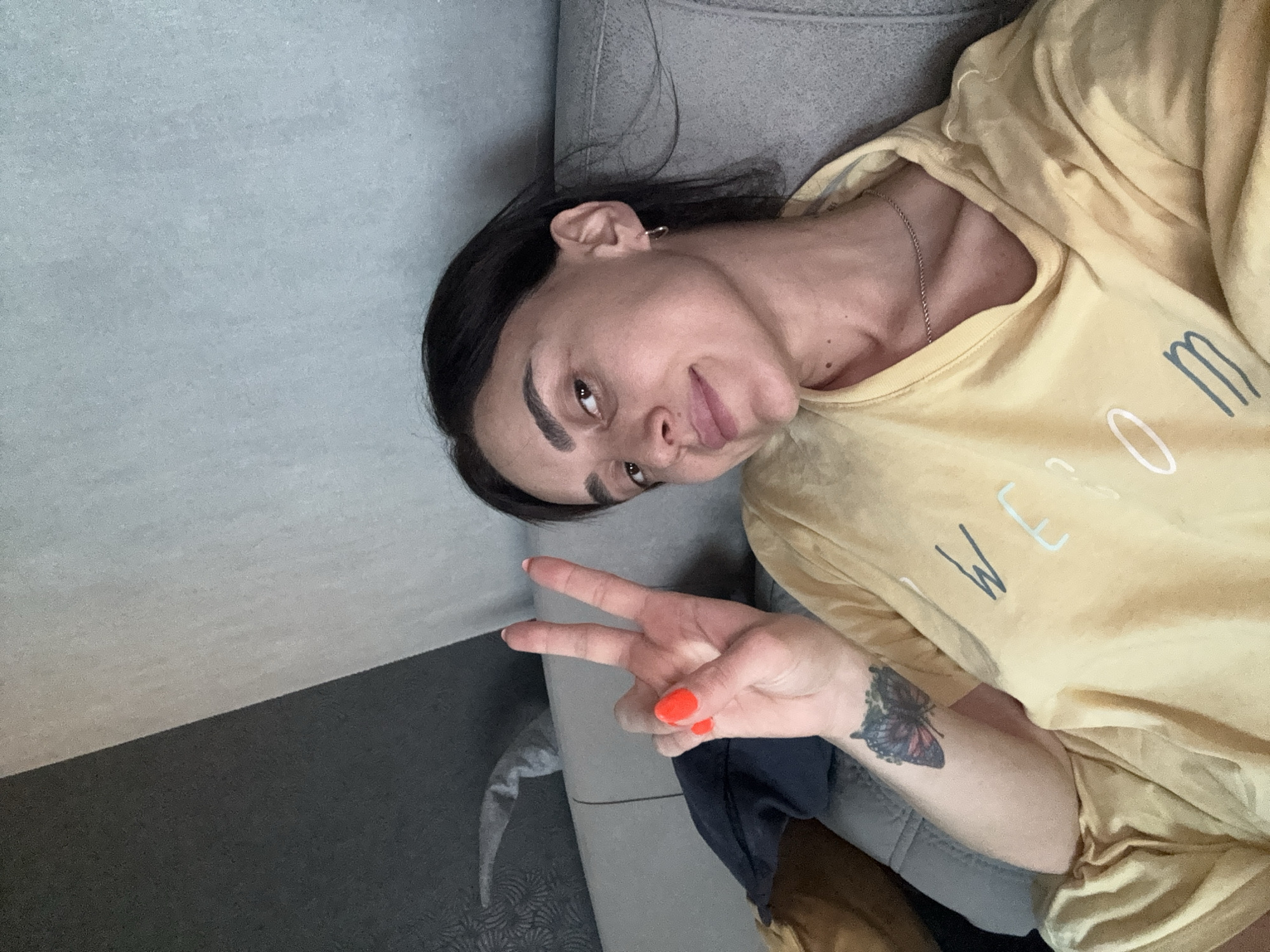 A person is lying on a grey couch, taking a selfie. They have dark hair, dark eyebrows, and brown eyes. They are wearing a yellow t-shirt with white and blue lettering that reads "WE GO". They are holding up two fingers in a peace sign, with bright orange nail polish on their fingers. On their left forearm, there is a tattoo of a butterfly. The room appears to be dimly lit, suggesting it might be nighttime or a cloudy day. The overall mood seems relaxed and casual. The person is looking directly at the camera with a slight smile. The couch appears to be in a domestic setting, possibly a living room. The background is a mix of grey wall and couch.