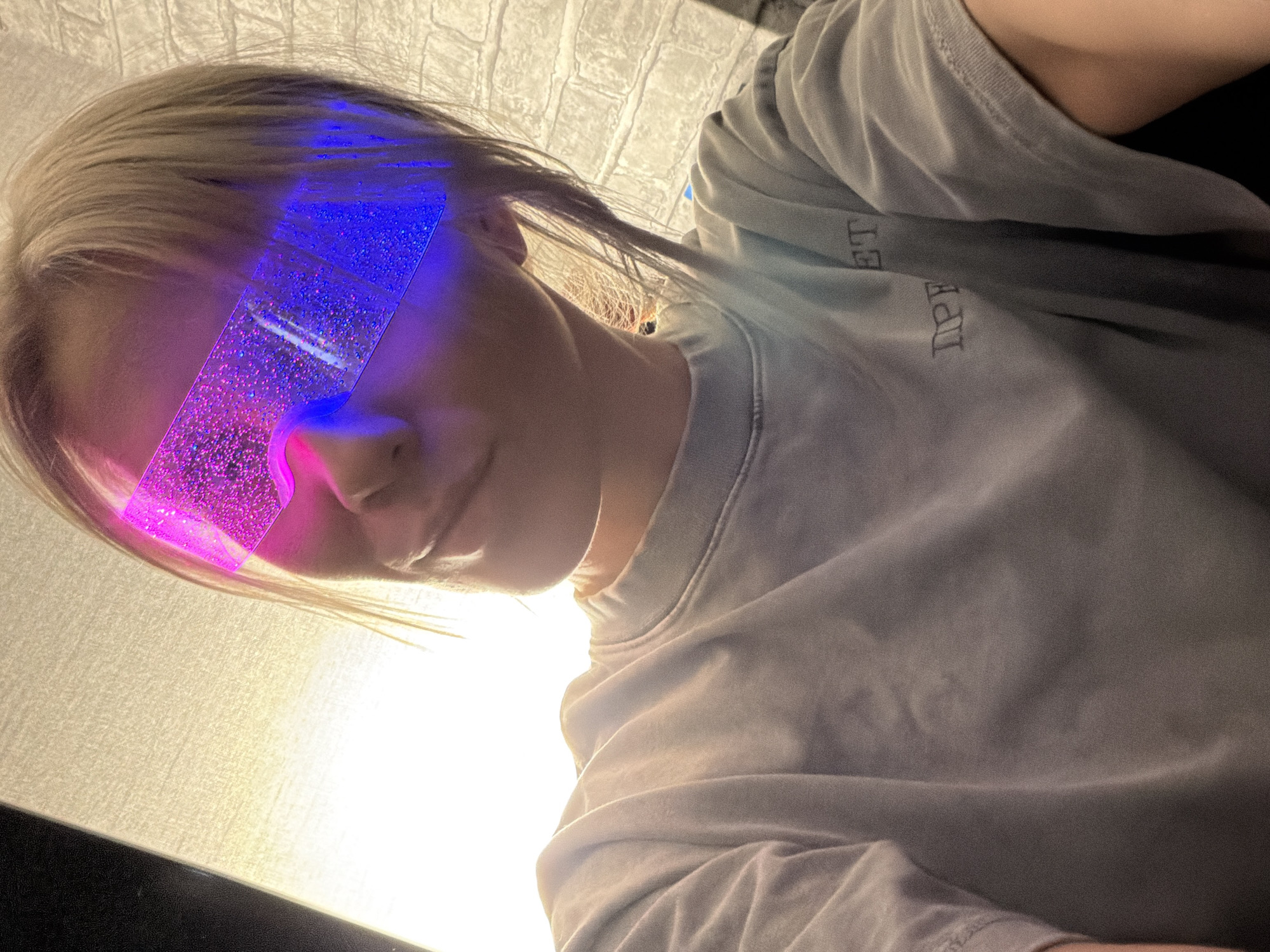 A person with blonde hair is wearing what appear to be novelty glasses that emit blue and pink light. The person is wearing a gray t-shirt with some text that is partially obscured. The background appears to be a wall with a textured pattern. There is no indication in the image that can identify the location as Chaikovsky, Russia.