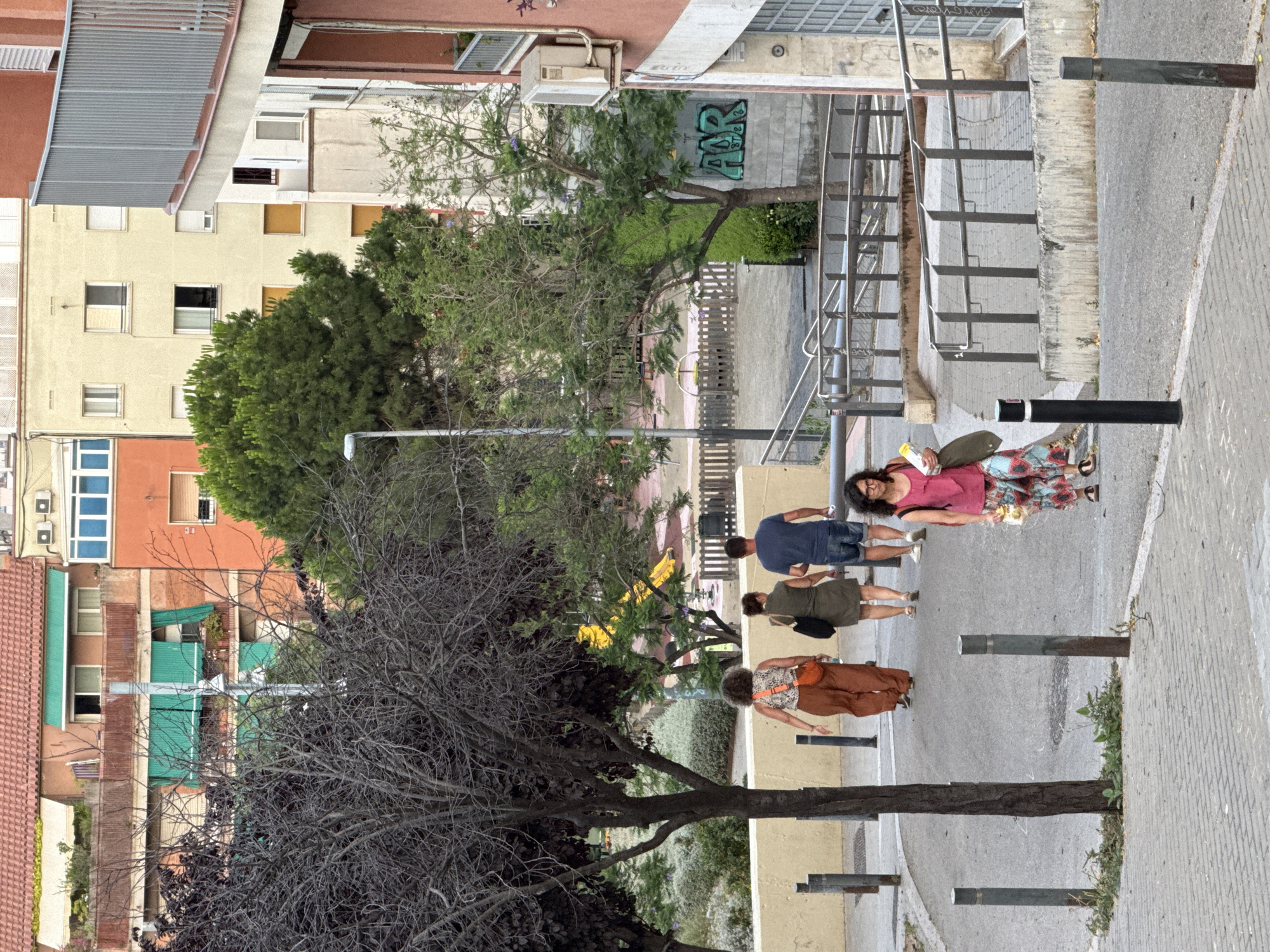 In a sun-drenched Barcelona street, four individuals walk down a paved sidewalk lined with metal bollards. The first person, a woman in a pink sleeveless top and a floral skirt, carries a small bag. She is followed by a man in a blue t-shirt and denim shorts, then a woman in a green dress with a black purse, and finally a woman in a patterned top and brown pants, also carrying a purse. The street is flanked by multi-story buildings with balconies and windows, and lush green trees line the sidewalk. The scene is bathed in natural light, suggesting it is daytime, with a clear sky. A metal staircase with handrails ascends on the right side of the image. Graffiti art in teal spray paint adorns a concrete wall in the background. The overall atmosphere appears casual and relaxed, as people stroll through the urban environment.