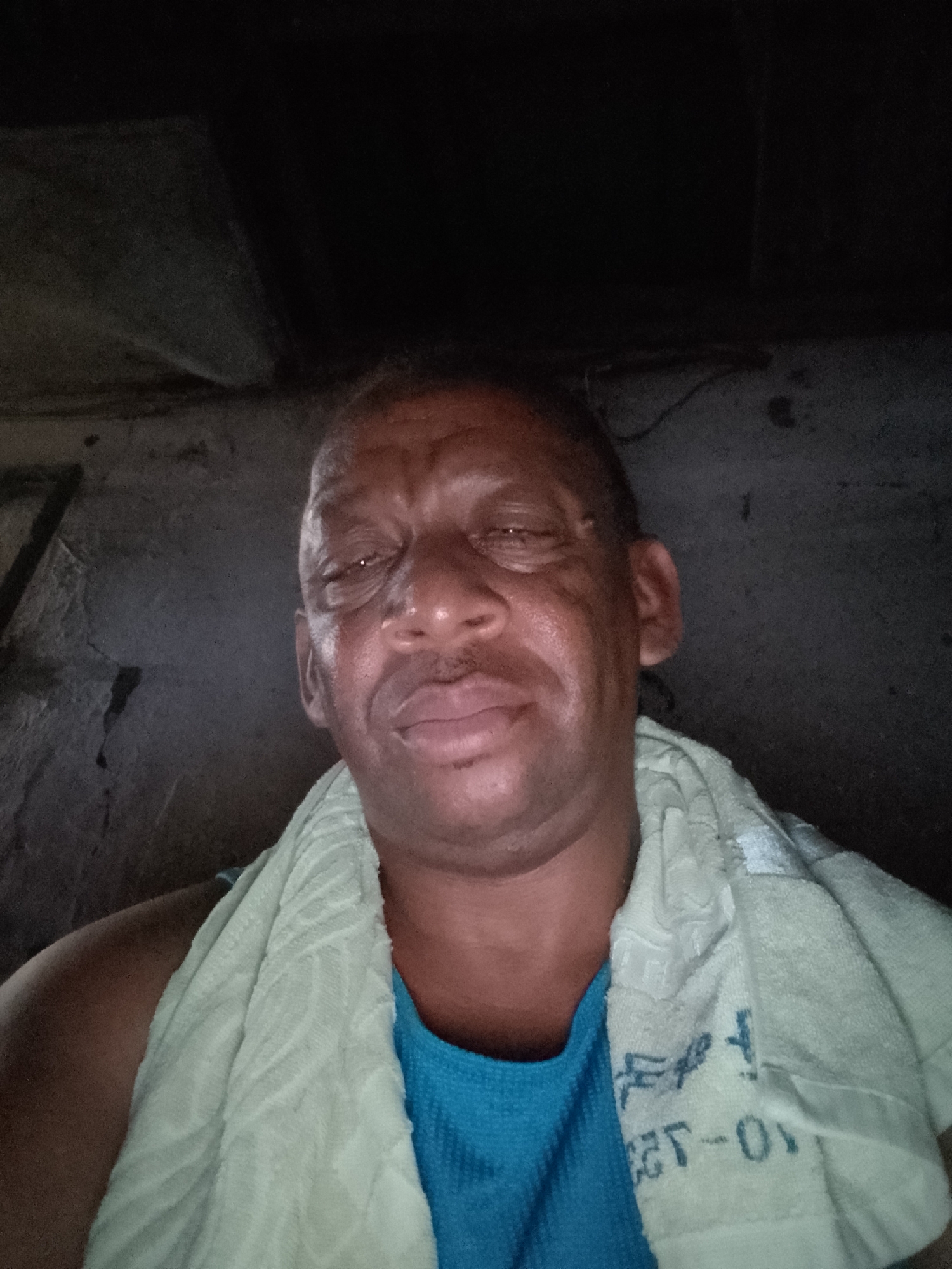 A close-up self-portrait of a dark-skinned man wearing a blue shirt and a light-colored towel around his neck. He has close-cropped hair and appears to be indoors. The background is dimly lit, showing a rough wall and a dark ceiling. There's no specific information to confirm it was taken in Sabon Tasha, Nigeria.