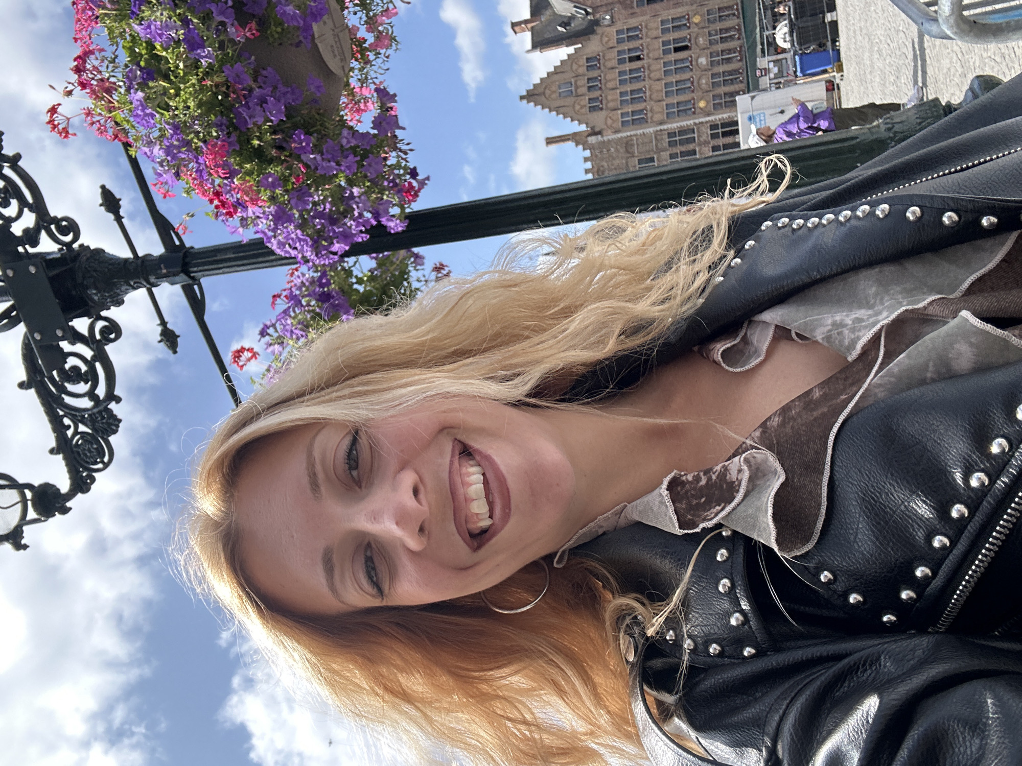 A woman with blonde hair smiles at the camera in a selfie taken outdoors. She is wearing a black leather jacket with silver stud details and a ruffled shirt underneath. Behind her, a lamppost with hanging baskets of purple and pink flowers is visible, along with a row of traditional Flemish-style buildings in Bruges, Belgium. The sky is blue with scattered clouds.