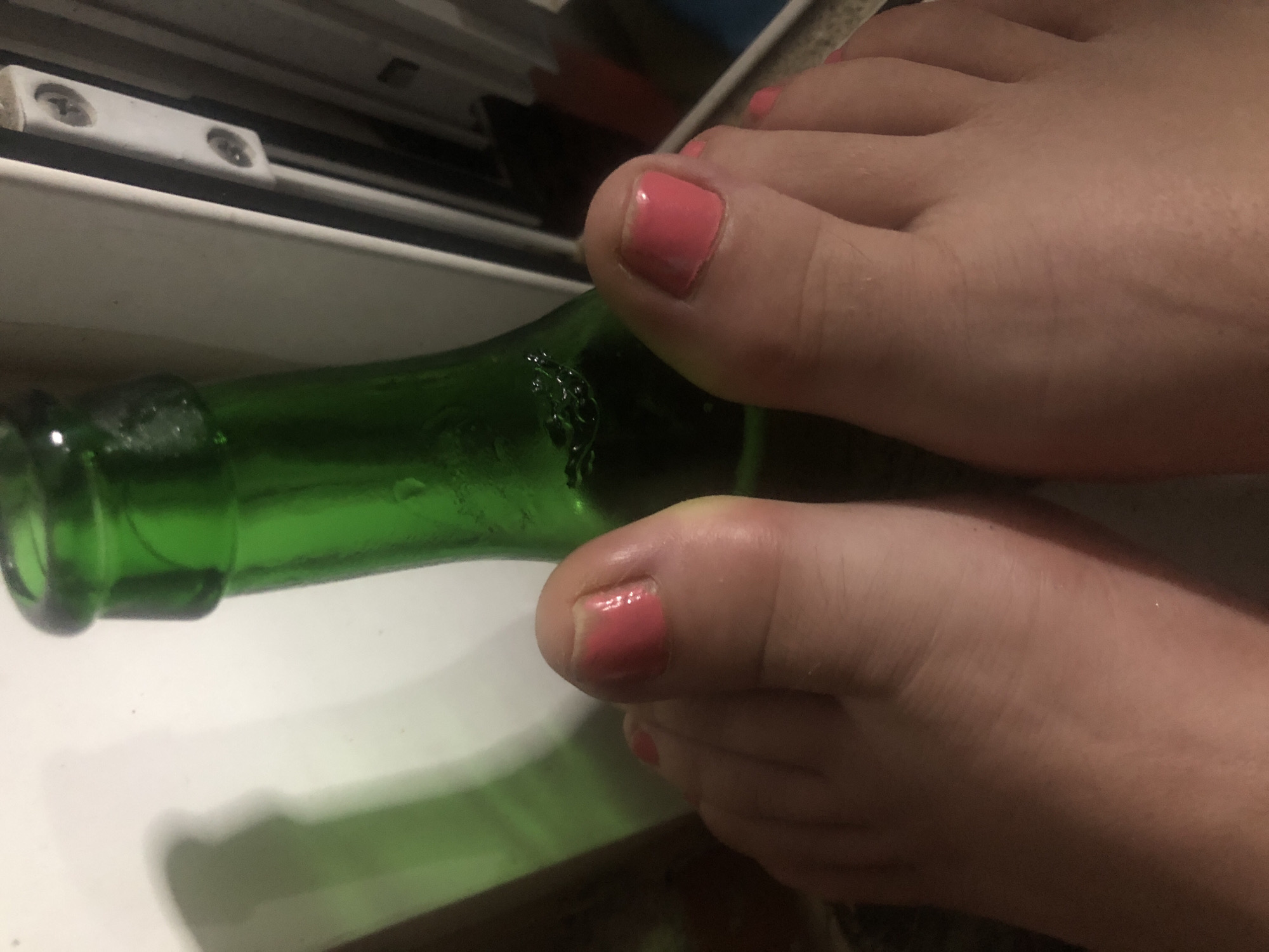 A close-up, eye-level shot shows a person's feet in the foreground, with a green glass bottle positioned behind them. The feet are bare, and the toenails are painted with a bright pink polish. The bottle is partially obscuring the background, which appears to be a white surface, possibly a wall or a refrigerator, with a light fixture and some screws visible on the right side. The lighting suggests an indoor environment. The specific location in the city of Unknown is not determinable from the image.