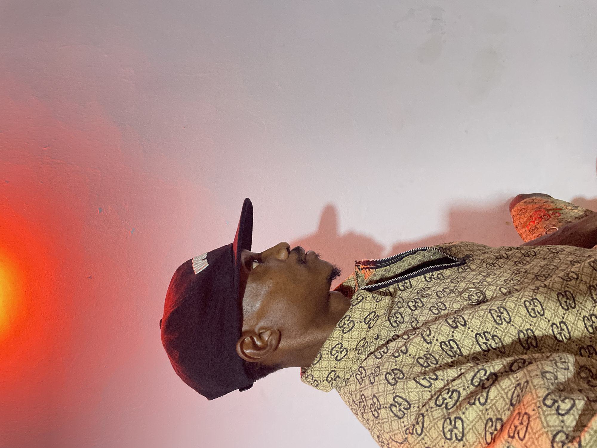 A person, identified as a male with dark skin and a short beard, is pictured in a side profile facing right. He is wearing a black baseball cap with white lettering and a light-colored jacket adorned with a repeating brown pattern of what appears to be the Gucci logo. The background is a textured white wall illuminated by a bright red light source from above. The scene is set in Adankolo, Nigeria.