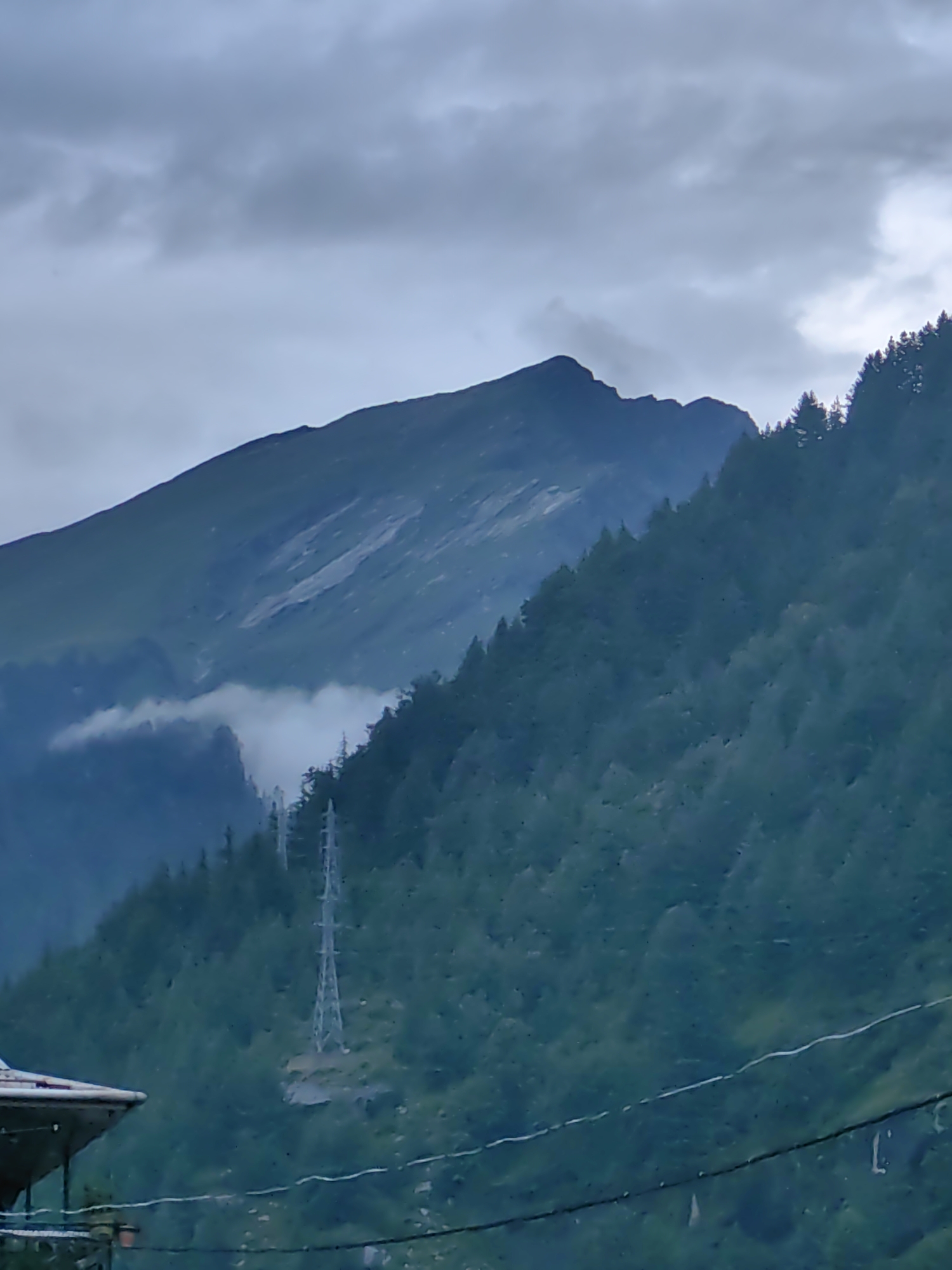 This media file captures a mountain landscape under a cloudy sky. The prominent subjects are a large, dark mountain range dominating the background and a densely forested mountainside in the mid-ground. A tall, lattice-like power line tower stands out on the slope of the forested mountain. In the foreground, the roof of a building is partially visible on the left, with some wires running horizontally across the frame.
The scene appears to be set during the daytime, with the overcast sky suggesting a possibly cool or rainy weather condition. The presence of clouds and the muted, cool tones of the image evoke a somber or serene mood. The location cues, such as the power line tower and the style of the building in the foreground, along with the type of mountain vegetation, might suggest a mountainous region in India, possibly in a remote or rural area. There are no visible people or specific activities depicted, presenting a static natural scene with man-made infrastructure. No text is visible in the media file.