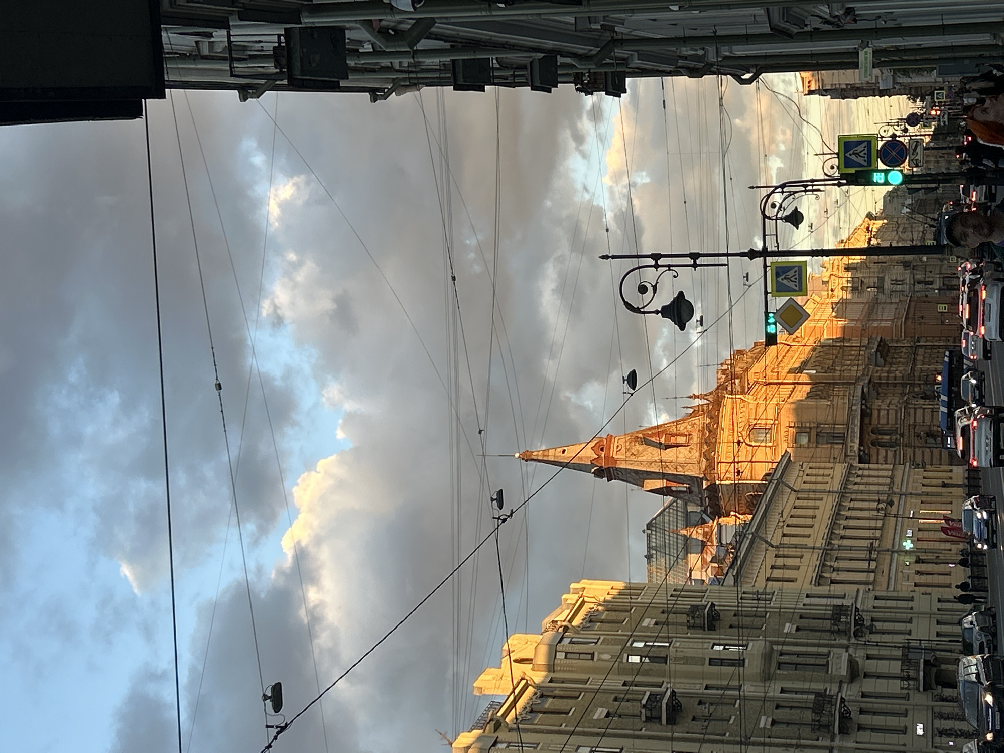 A view down a city street at dusk, with tall, ornate buildings lining both sides of the road. The sky is filled with dramatic, gray clouds highlighted by the warm glow of the setting sun. Overhead, a complex network of wires crisscrosses the scene, along with several streetlights. Cars are visible driving on the street, and a few pedestrians can be seen on the sidewalks. A prominent, pointed spire of a building catches the golden light, dominating the center-left of the frame. Traffic signals are present, with one showing a green light. The architecture of the buildings suggests a European city.