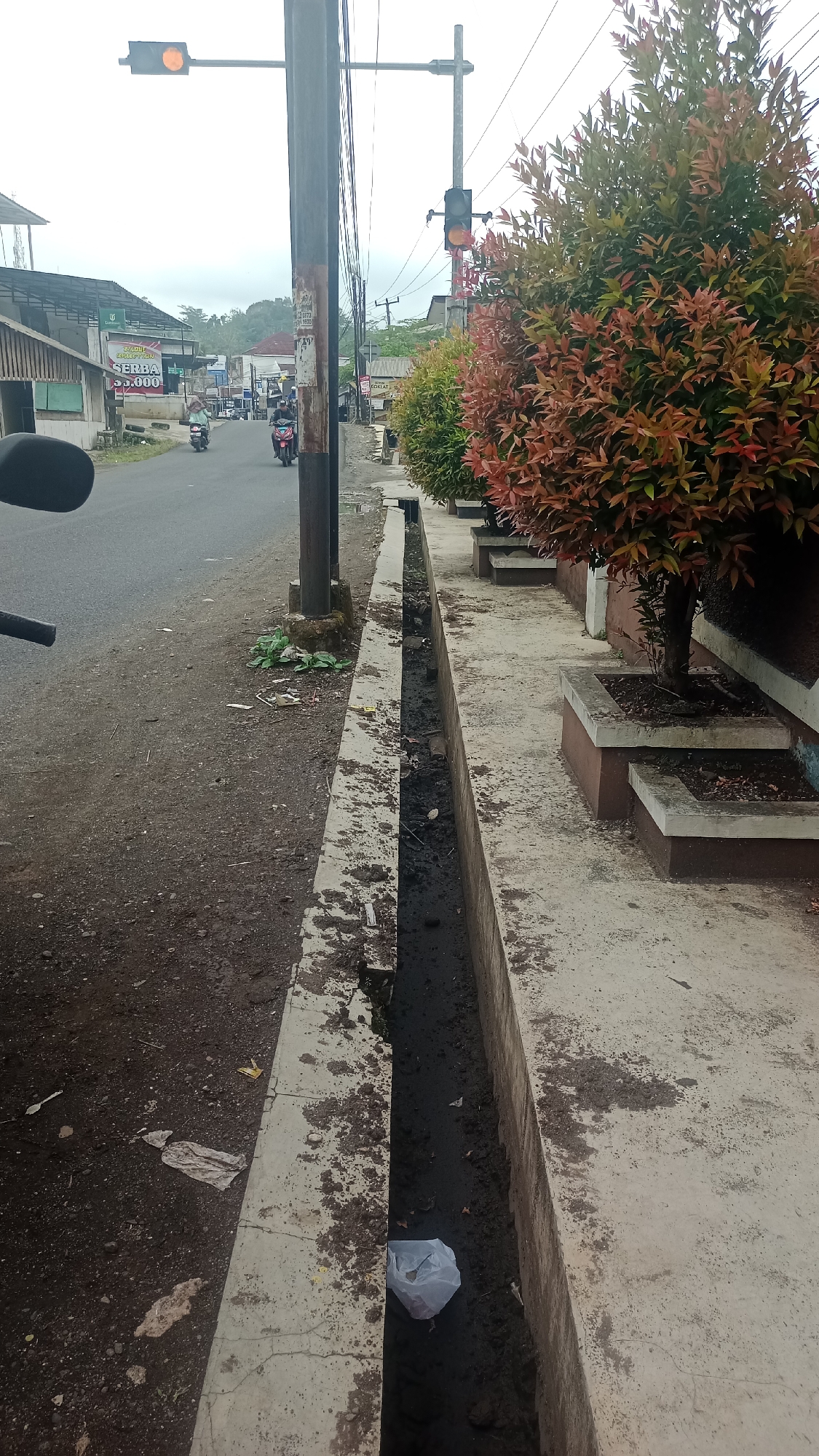 The image depicts a street scene in Karyamandala, Indonesia. On the left side of the frame, a paved road is visible, with a grey motorcycle in the foreground and two motorcycles with riders further down the road. A traffic light with a yellow signal is mounted on a pole above the road. To the right of the road is a concrete drainage ditch filled with dark, stagnant water and some debris, including a white plastic bag. A concrete sidewalk runs alongside the ditch, and beyond that, a row of potted plants with vibrant red and green foliage lines the walkway. In the background, buildings and trees line the street, suggesting a commercial or residential area. The sky is overcast, contributing to the muted lighting of the scene.