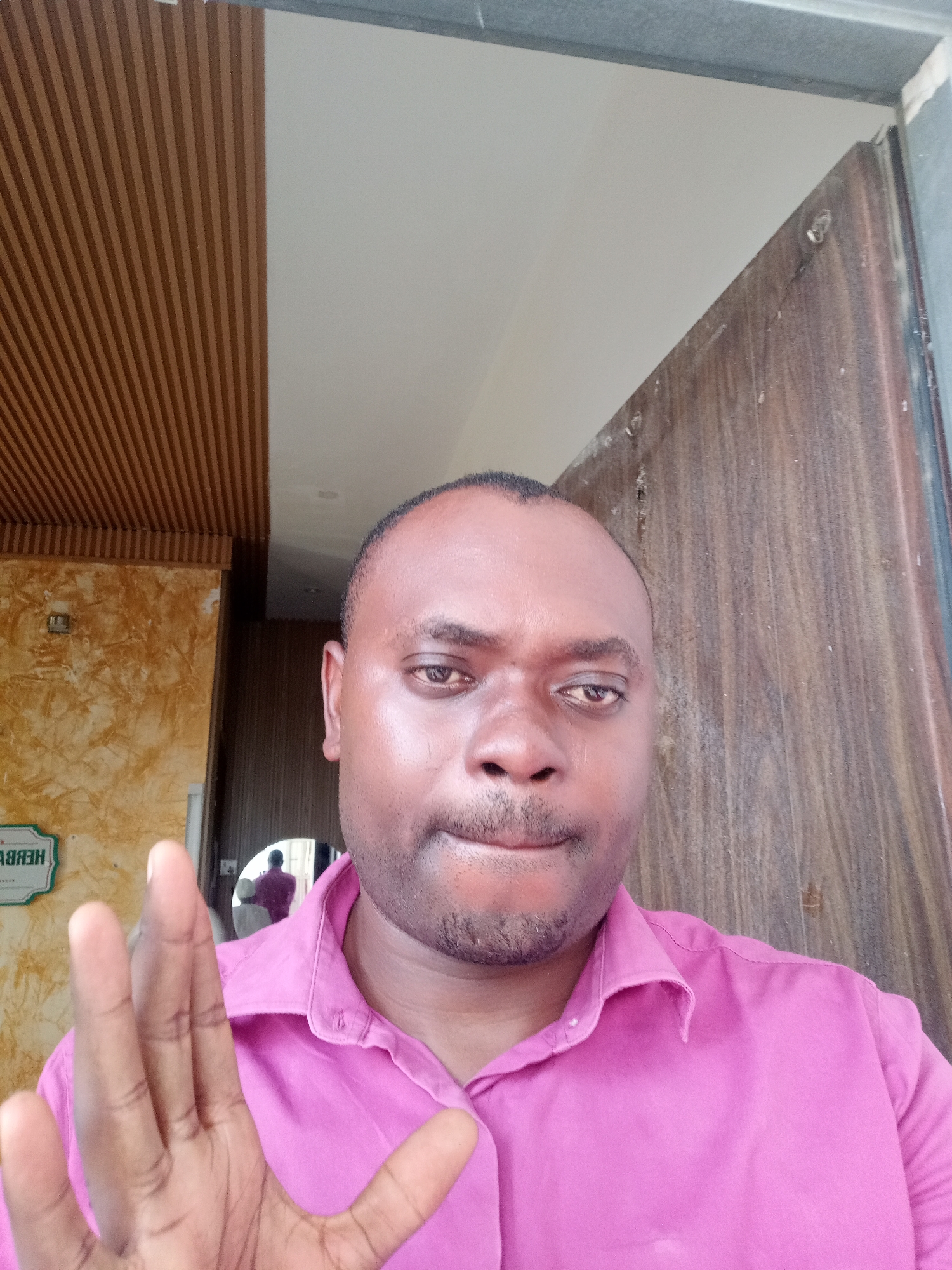 A man with dark skin, short black hair, and wearing a pink collared shirt is taking a selfie. He is extending his left hand towards the camera. In the background, there is a wooden door and a paneled wall. The location appears to be indoors in Gwarinpa, Nigeria. A reflection of a figure in a red shirt is visible in a mirror. A sign with illegible text is partially visible.