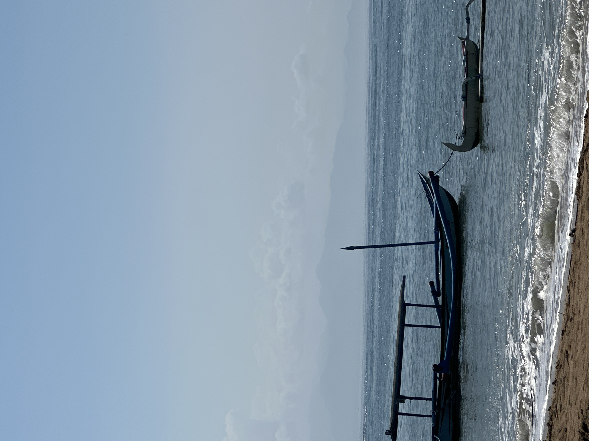 Two boats are anchored in the calm ocean waters off the coast of Denpasar, Indonesia. The larger boat, painted blue, is closer to the shore and features a canvas roof supported by wooden poles. A thin, dark pole with a pointed tip extends vertically from the water near the boat. A smaller, lighter-colored boat with a distinctive curved prow is tied to the larger boat by a rope and floats further out. The water exhibits gentle waves that break onto a sandy beach in the foreground. In the distance, misty mountains are visible, partially obscured by haze. The sky is a pale blue, with faint clouds towards the horizon. No people are visible in the frame.
