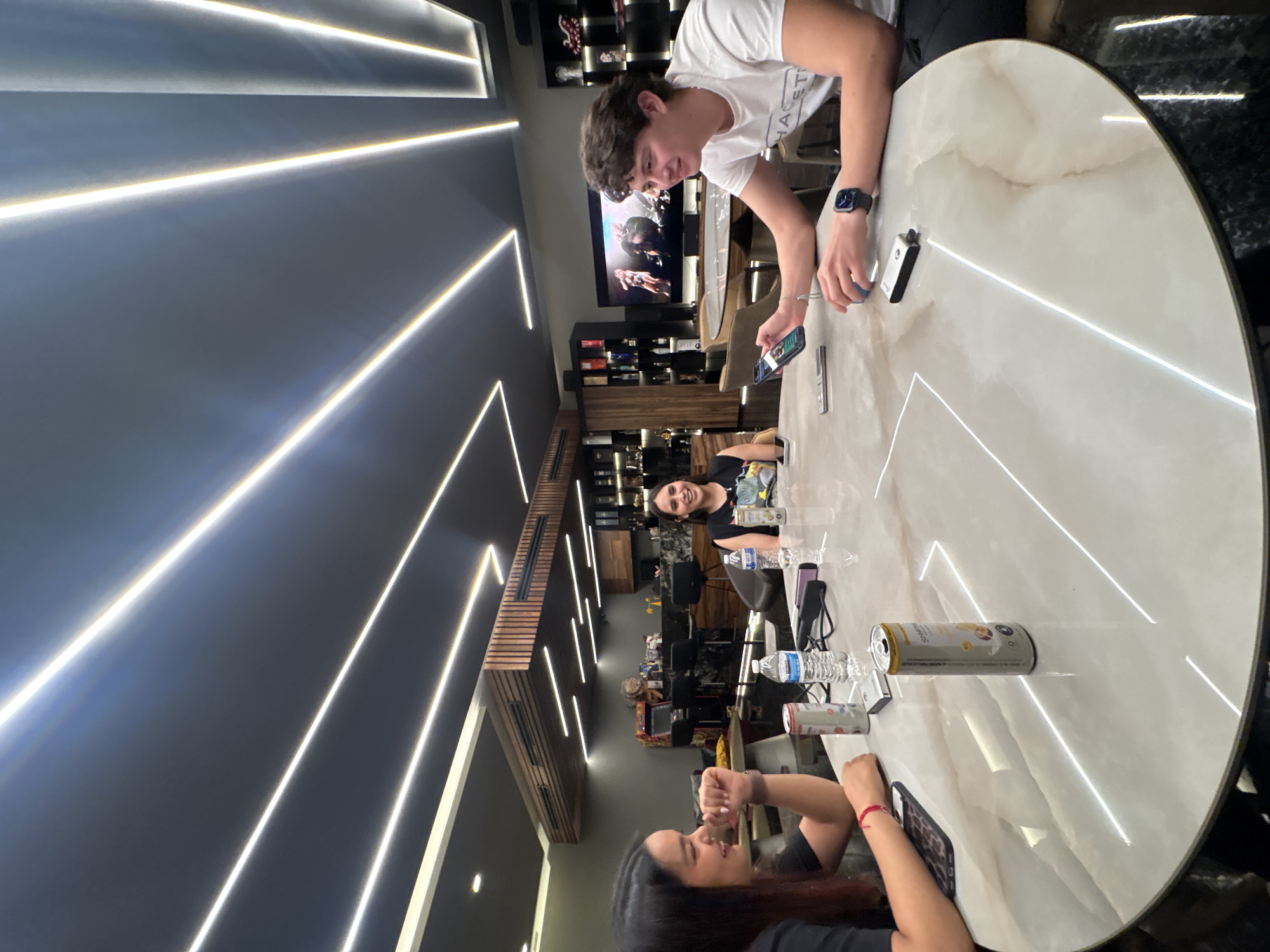 The image shows a group of people sitting around a large, round table in a well-lit interior. The table is marble-like, with embedded linear lights. On the table are cellphones, drinks, a water bottle, and a vaping device.
One person, a young man wearing a white T-shirt, is standing at the edge of the table and looking down at a phone in his hand. Two women are seated at the table. One is smiling towards the viewer.
In the background is a wall-mounted TV and display shelves filled with bottles. Above is a modern ceiling with integrated linear lights. The exact location within San Bernardino Tlaxcalancingo, Mexico is not possible to determine from the image alone.