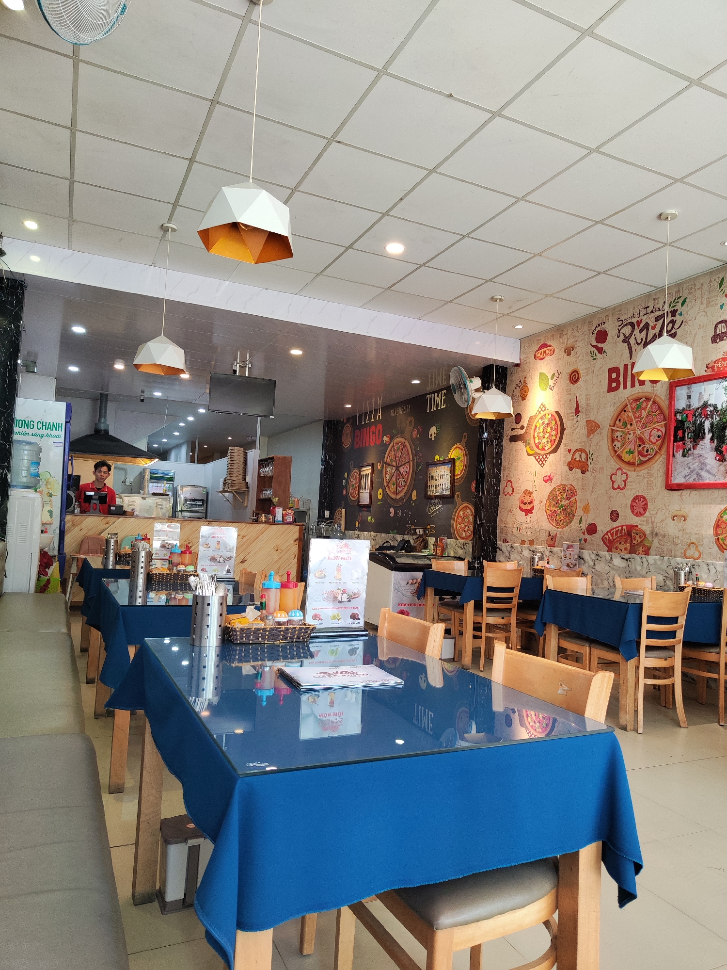 The interior of a restaurant in Da Lat, Vietnam is visible. Tables with blue tablecloths and wooden chairs are arranged throughout the space. Hanging lights with white shades and orange accents illuminate the area. One worker can be seen behind a counter. The walls feature pizza-themed artwork and signage, including the words "Pizza Bingo". A water dispenser and other food service items are present.