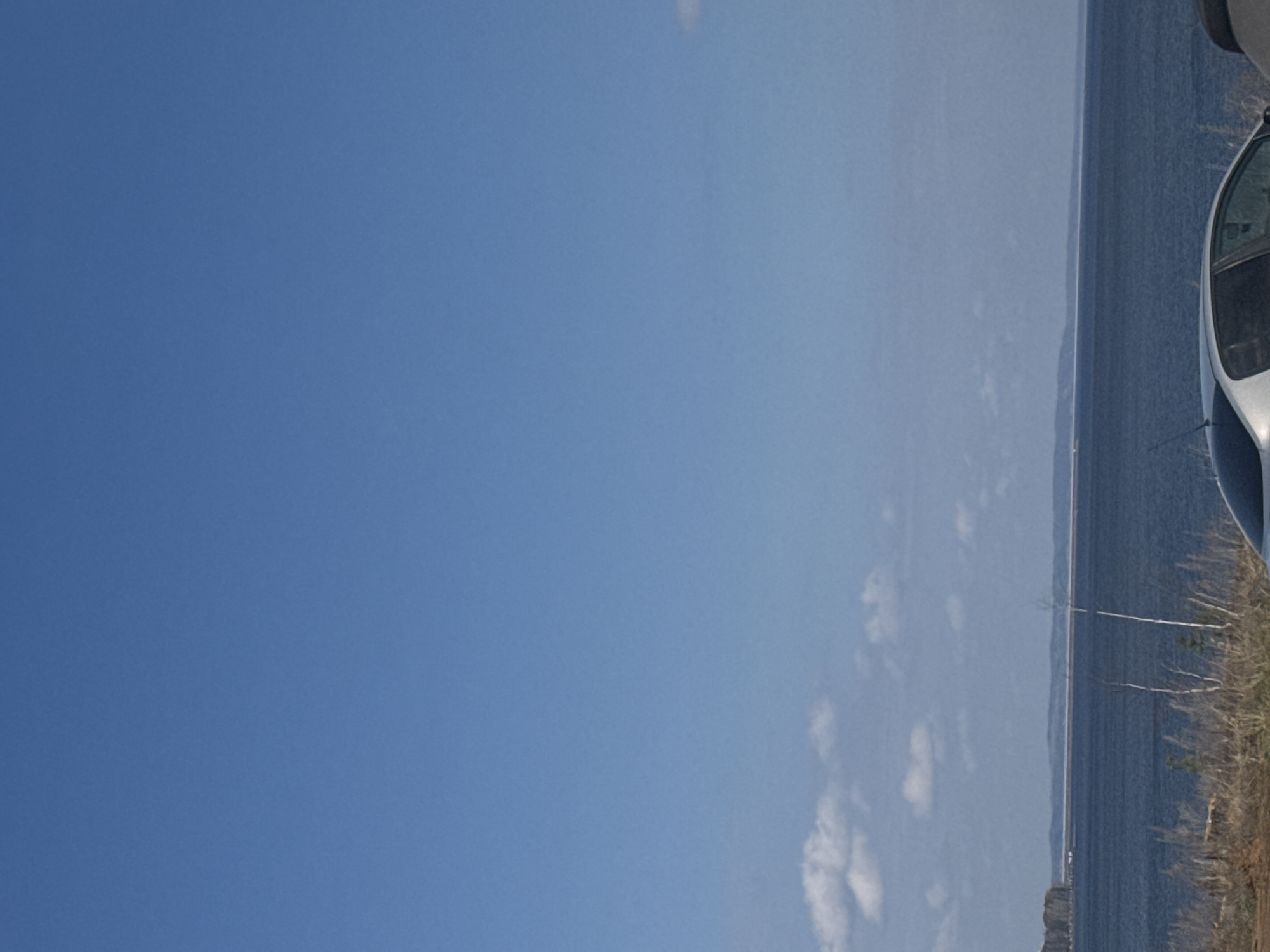 The image shows a view of the Kama River in Chaikovsky, Russia. The sky is clear and blue, taking up the majority of the frame. A body of water, the Kama River, is on the right-hand side with a thin strip of land visible on the far bank. Some light vegetation and what appears to be the side of a car are visible on the close right edge of the image.