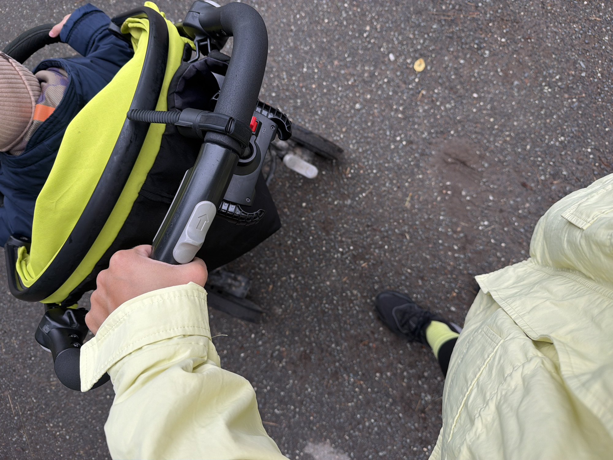 This top-down, first-person perspective image captures an adult pushing a child in a tricycle-stroller hybrid on a paved outdoor surface in Votkinsk, Russia.
**People:**
* An adult, likely the photographer, is visible from the chest down. The right hand firmly grips the left side of the vehicle's handlebar. The adult wears a light yellow or lime green crinkled jacket or raincoat with visible stitching and a pocket outline on the left side of the torso. Black leggings or pants are worn, paired with bright neon yellow-green socks and black athletic shoes with white accents.
* A child is seated in the front portion of the vehicle, facing away from the photographer. The child wears a light beige knit beanie, a dark blue jacket, and a patterned scarf or neck gaiter in plaid-like purples and browns with a prominent orange stripe. The child's right hand is visible, seemingly gripping a part of the vehicle's frame.
**Subjects/Objects:**
* **Child's Vehicle:** A child's push tricycle or a stroller-tricycle hybrid is central to the image. Its frame is predominantly black. The child sits within a bright neon yellow-green fabric enclosure, likely a canopy or seat liner, which is trimmed with a thick black border.
* **Handlebar:** The adult's handle is black, featuring a textured grip. A grey plastic mechanism with a white arrow icon (indicating an adjustment point) is visible on the left side. A red circular button, likely a brake or lock, is present on the right side of the handlebar assembly. Black straps or ties are fastened around the handlebar, possibly securing an accessory.
* **Wheels/Pedal:** Partially visible wheels suggest a tricycle design. A white pedal or footrest is faintly discernible beneath the handlebar on the left side of the vehicle.
**Actions/Interactions:**
* An adult is actively pushing the child's vehicle.
* The child is passively riding in the vehicle, appearing to hold onto its frame.
**Environment:**
* The ground surface is a dark grey asphalt or paved path, characterized by small, embedded light-colored pebbles, indicating a granular texture. The surface appears slightly damp in areas, suggesting recent moisture.
* Minimal environmental debris is present, with one small, light yellowish, round leaf or seed pod visible on the ground in the lower right quadrant.
* The lighting is even and diffused, typical of an overcast sky, with no distinct shadows cast.