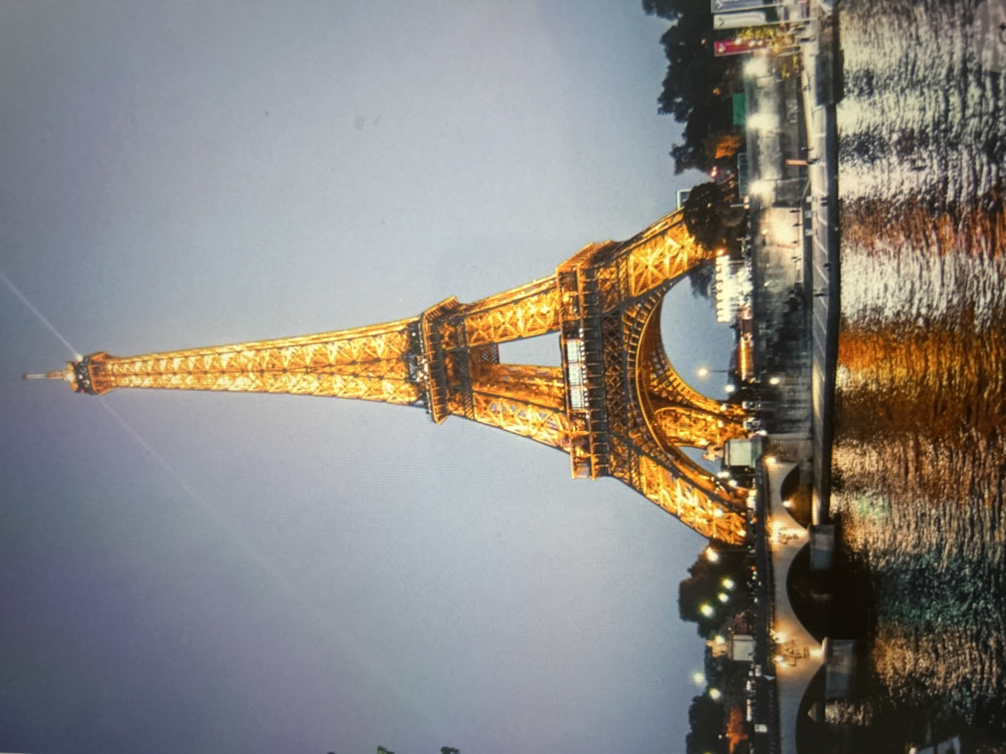 The image, rotated vertically, captures the illuminated Eiffel Tower at night, set against a dark, slightly overcast sky. The tower, a prominent architectural landmark, glows in a warm golden-orange hue, highlighting its intricate iron lattice structure. It stands tall on the left side of the frame, with its peak extending towards the top left corner.
To the right of the tower, a body of water, most likely the River Seine, is visible. The water's surface reflects the bright lights of the tower and other sources on the riverbanks and a nearby bridge, creating vibrant streaks and shimmering patterns. Along the riverbanks, various structures and silhouetted trees are visible, also lit by artificial lights. A bridge with arched supports spans the river in the foreground, featuring lights along its length that contribute to the strong reflections in the water.
Indistinct figures or shapes, possibly people or street furniture, can be seen along the riverbank and on the bridge, appearing as small, blurred forms against the luminous backdrop. The time of day is clearly evening or night, emphasized by the dark sky and widespread artificial illumination. The scene conveys a sense of grandeur and iconic beauty. There is no visible text that can be deciphered.