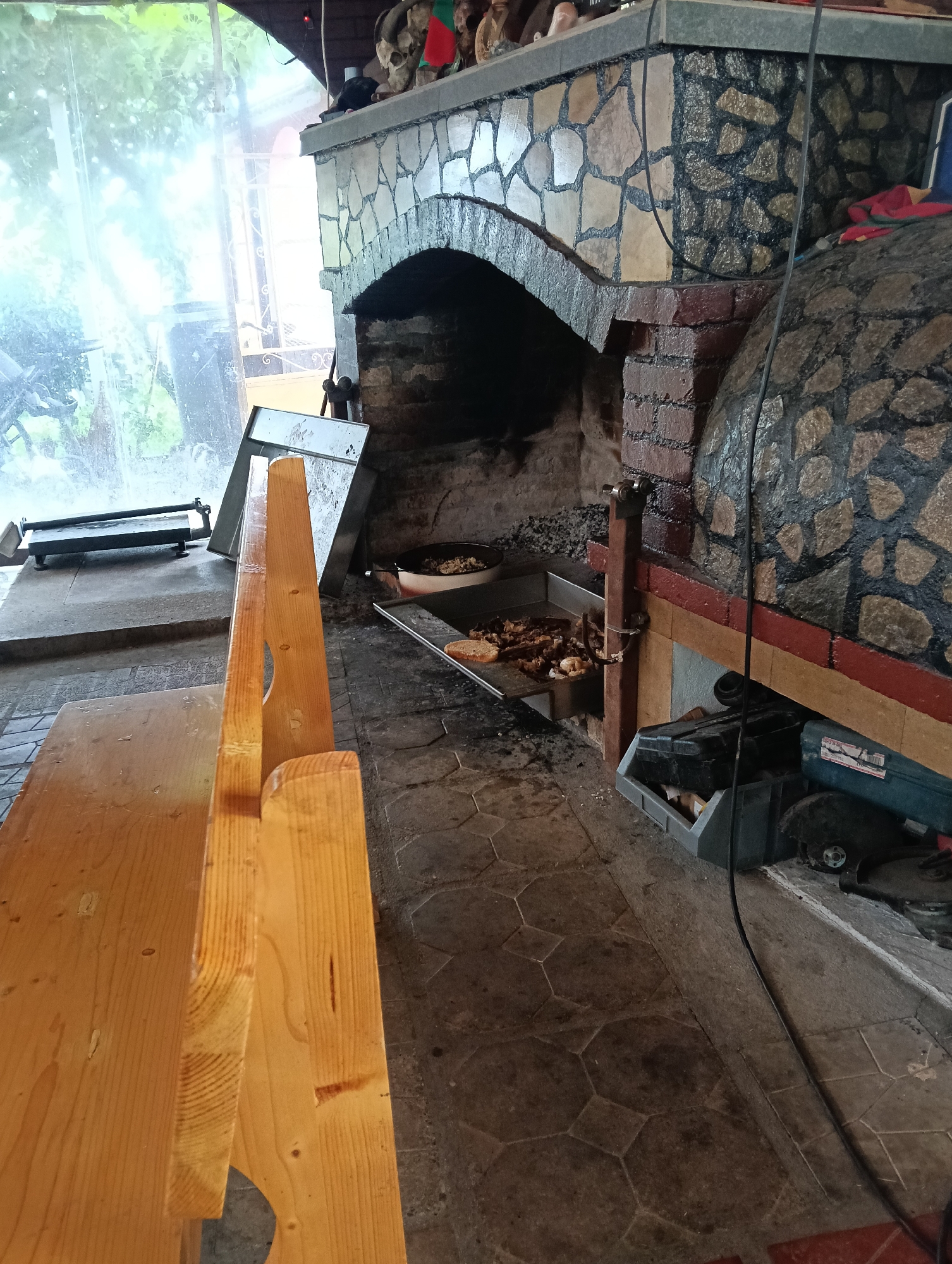 The image depicts an outdoor setting, likely a rustic outdoor kitchen or dining area, near a fireplace or oven. In the foreground, a simple wooden bench is positioned on a tiled floor. The bench is made of unfinished pine wood and appears to be a backless design.
Dominating the background is a large, stone-clad structure that resembles a fireplace or a pizza oven. The arched opening of the fireplace reveals an interior filled with dark coals or embers. A metal tray containing food, possibly grilled meats and bread, is placed in front of the fireplace. Next to the food tray, another metal tray is visible.
To the left of the fireplace, a clear plastic sheeting or curtain is seen, allowing diffused natural light to enter the space. Beyond the sheeting, some greenery and parts of outdoor furniture are faintly visible, suggesting a garden or patio setting.
On the right side of the image, near the base of the stone structure, several plastic storage containers and what appears to be a power tool are present. The flooring throughout the scene consists of irregularly shaped, hexagonal, and square tiles.
The overall impression is one of an informal, possibly rural, establishment where food is prepared and served outdoors. Based on the context, this scene is located in Konush, Bulgaria.