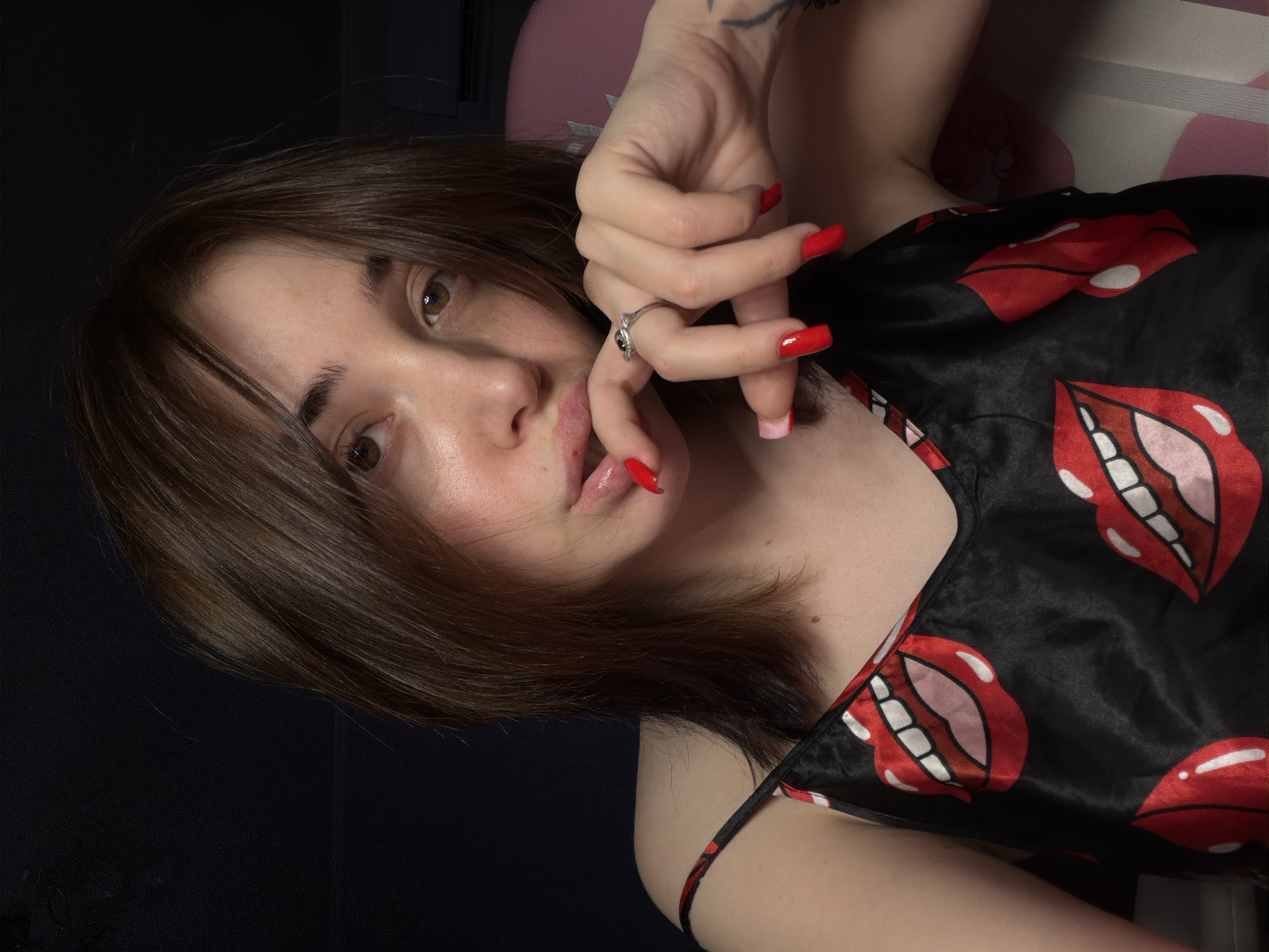 A close-up, chest-up, medium shot captures a young Caucasian woman with shoulder-length brown hair and light brown eyes. She is looking at the camera, with her right hand resting on her mouth, her index finger touching her lips. Her nails are painted a vibrant red. She is wearing a black silk camisole with a bold red and white lips pattern. A silver ring with a dark stone is visible on her left ring finger. The background is dark, with hints of a pink chair and a portion of a white air conditioning unit on the right side. The lighting is dim, casting shadows on her face and creating a mood of intimacy.