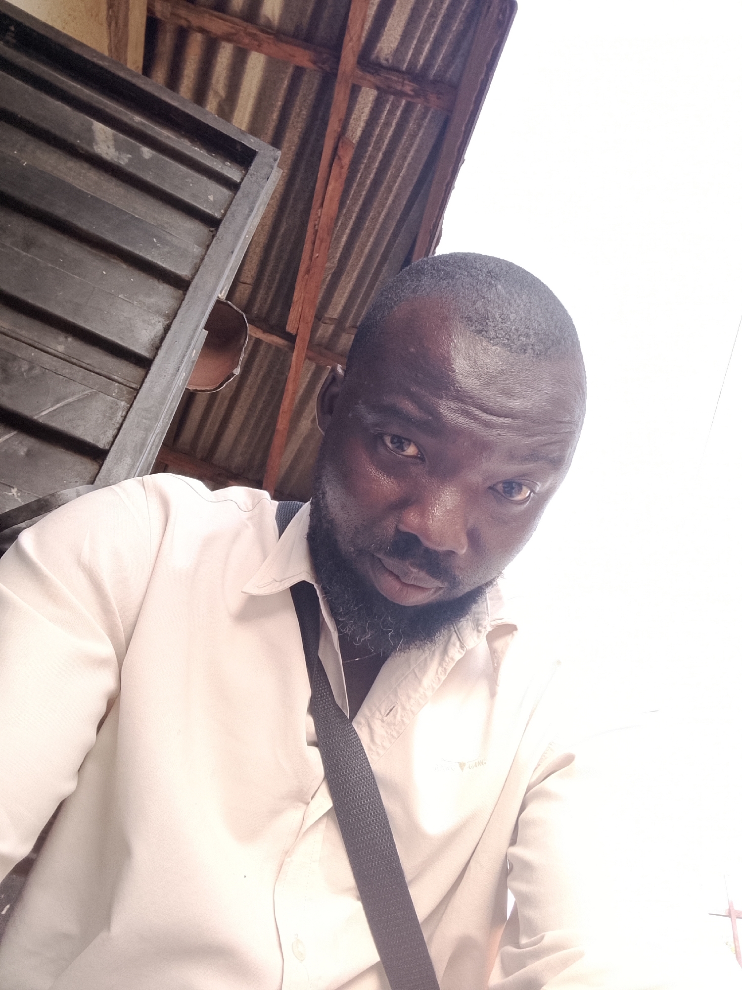 A medium-close shot shows a Black man with a shaved head and a neatly trimmed beard, wearing a light-colored button-down shirt and a dark strap, possibly from a bag, across his torso. His eyes are visible. The environment consists of the underside of a corrugated metal and wooden structure with a grey metal frame, likely a roof or awning. The location is likely Gwong, Nigeria. The man is the primary subject.