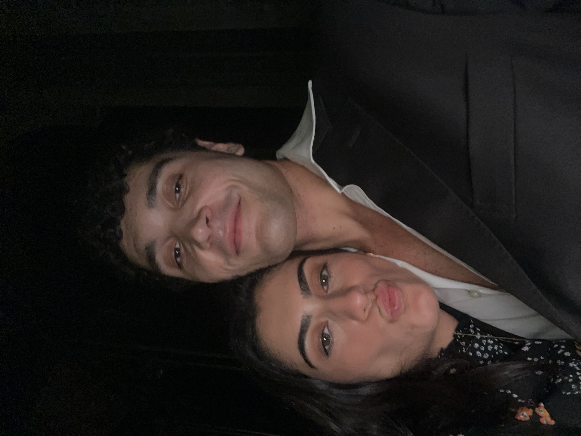 A selfie captured in close proximity, featuring a man and a woman against a dark, out-of-focus background. The woman is positioned to the left, leaning her cheek against the man's shoulder. She has long, dark hair, and her lips are pursed in a kiss pose. She wears a dark, floral-patterned top. The man, to the right, is looking forward with a slight smile and has curly dark hair. He is wearing a white collared shirt, partially unbuttoned, and a black suit jacket. The lighting appears to be low, with a warm tone illuminating their faces. There is no identifiable information suggesting a location in Paris, France.