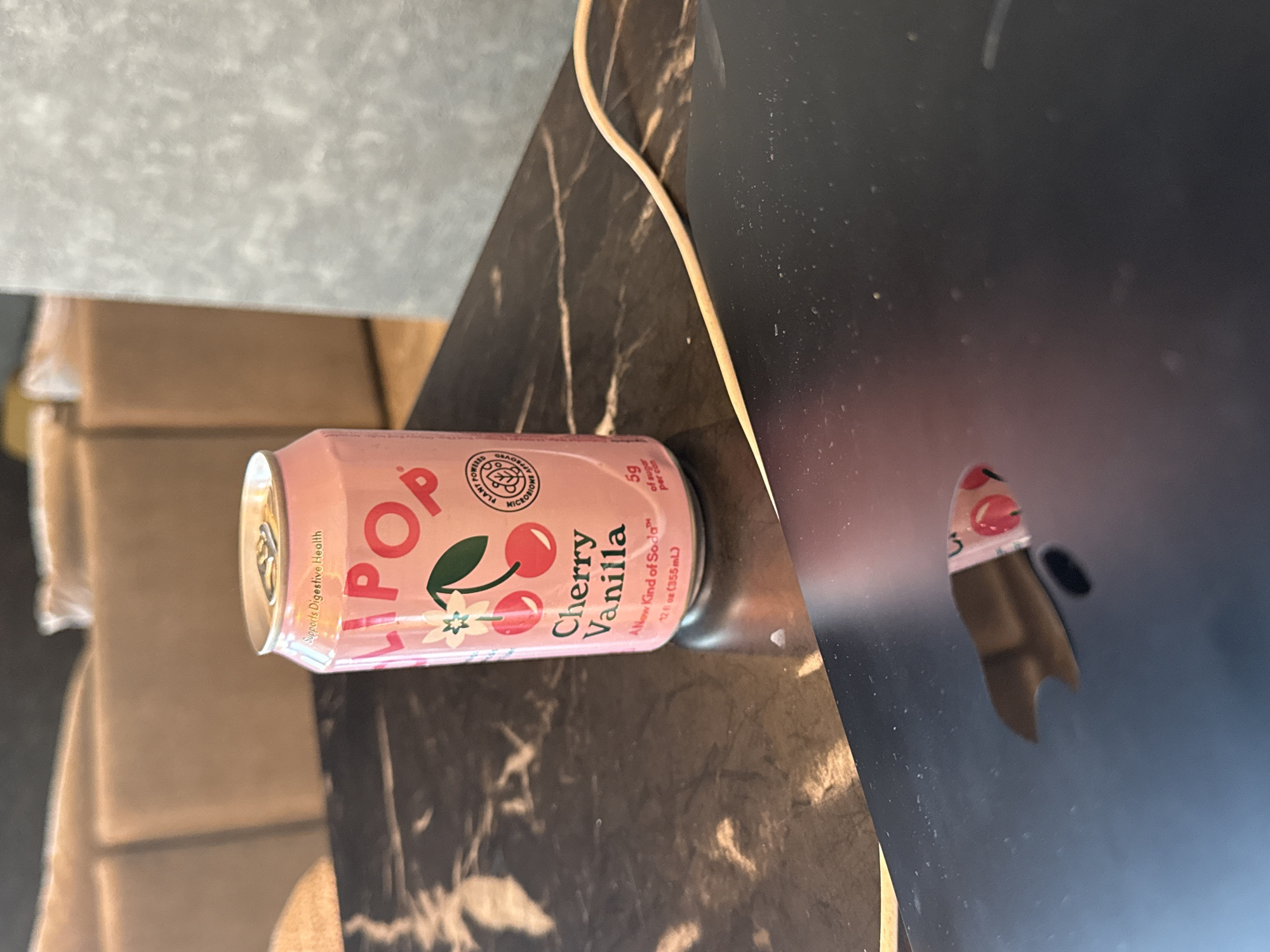 An indoor, medium-shot image features a pink OLIPOP soda can, specifically the Cherry Vanilla flavor, lying on its side on a dark, marble-patterned table surface. The can is positioned centrally in the frame, angled slightly downwards from left to right.
The can's label is prominently visible, displaying the brand name "OLIPOP" in large red letters. Below this, the flavor "Cherry Vanilla" is printed. The can features illustrations of cherries and small white flowers on its pink background. Several claims and details are visible on the label, including "Supports Digestive Health," "PLANT POWERED MICROBIOME APPROVED," and "A New Kind of Soda™." Nutritional information states "5g of sugar per can," and the volume is "12 fl oz (355mL)." The pop-top of the can is visible at the top left, showing it is unopened.
A light-colored wire runs diagonally across the table surface, partially obscuring the top right portion of the can. The dark table surface exhibits subtle reflections, including a distorted, reddish reflection of the can to the right. In the blurred background on the left, stacks of light brown cardboard boxes are visible. The upper background shows a textured, light gray surface, while the right side of the background reveals a dark, somewhat dusty surface, possibly part of another object or wall. The scene is evenly lit, indicating an indoor setting, likely under artificial lighting.