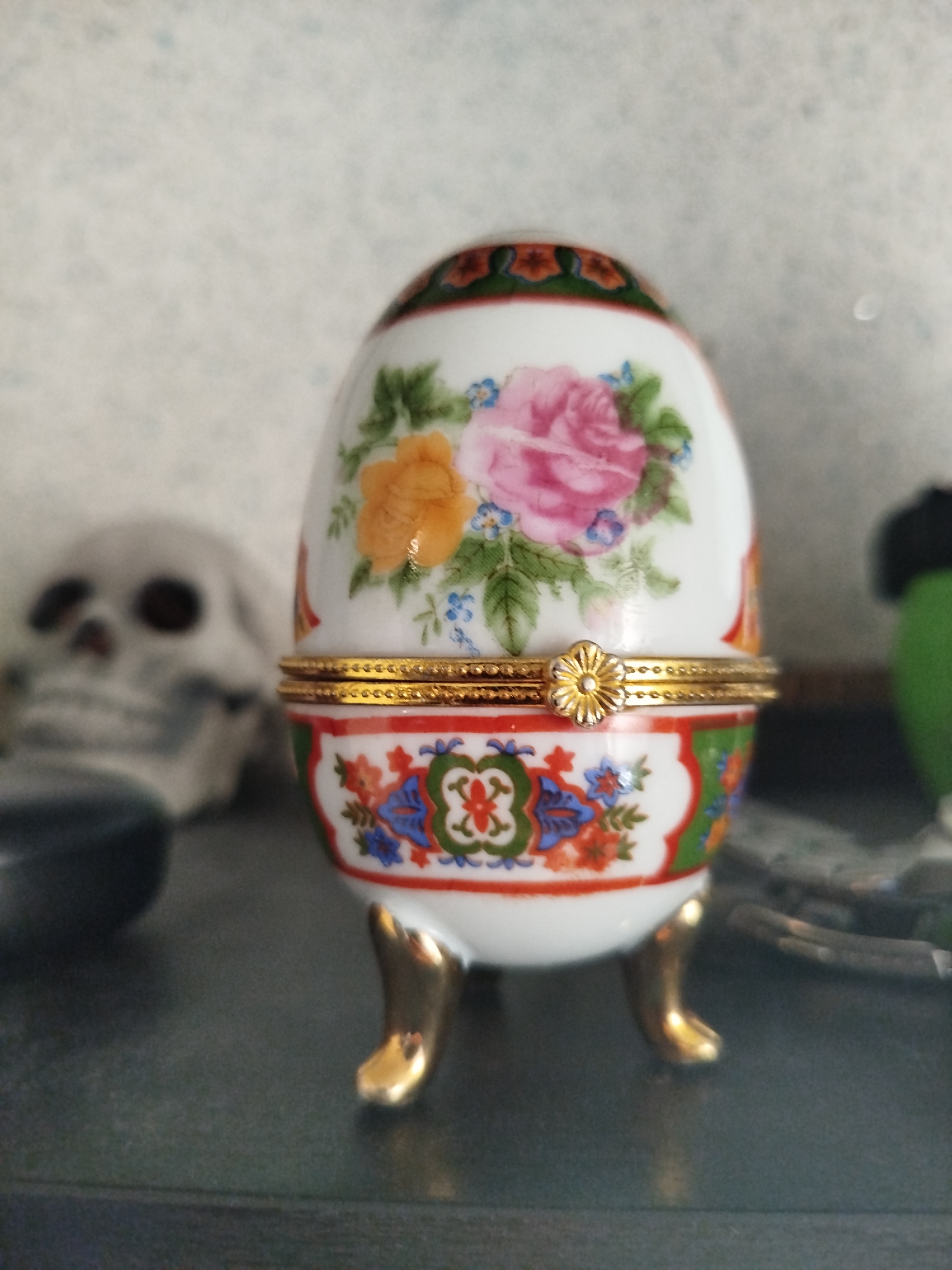 A decorative porcelain egg sits on a dark surface. The egg is white and adorned with painted pink and yellow roses and small blue flowers. A gold trim encircles the middle of the egg, featuring a small, flower-shaped clasp. The egg rests on four gold-colored legs. In the background, a blurry, light-colored object with dark eye sockets, resembling a skull, is visible to the left of the egg. A dark, blurry object is also present to the left, and a blurred green object is to the right. The background appears to be a textured, light-colored wall.