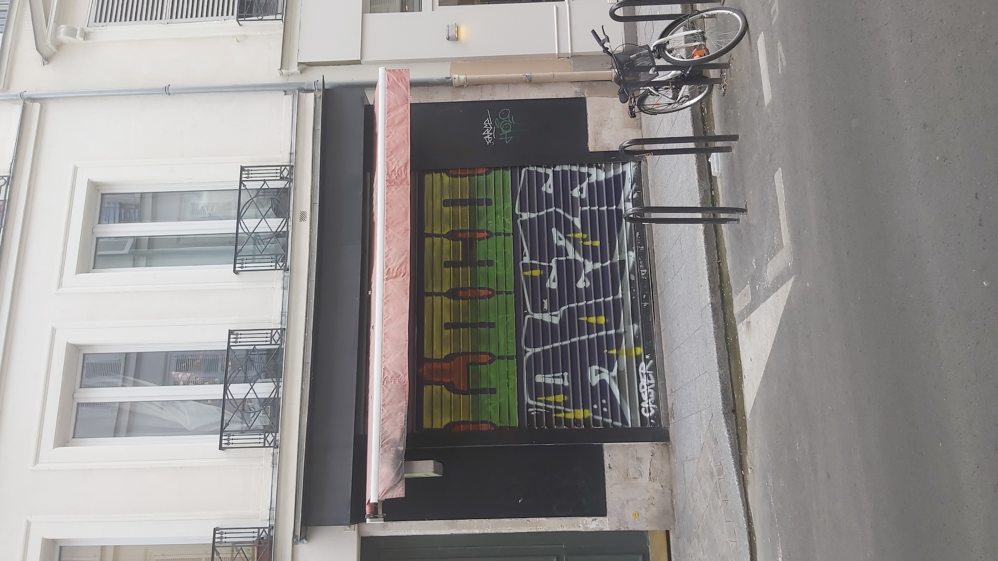 A roller-shutter door of a building in Paris, France, is covered in graffiti. The roller shutter is a dark grey color, with vertical lines indicating its segmented nature. The graffiti consists of two layers. The upper layer features large, stylized letters in orange, yellow, and green, with a gradient effect. The lower layer is dominated by graffiti in white and yellow, with thick, blocky letters. Below the main graffiti, a smaller tag in white reads "CASPER". The roller shutter is part of a storefront, with a light brown awning extended above it. The building facade is white, with two windows visible on the floor above. In front of the roller shutter, there are two metal bicycle racks. To the right of the racks, a white bicycle with a basket is parked. The street in front of the building is paved with asphalt.