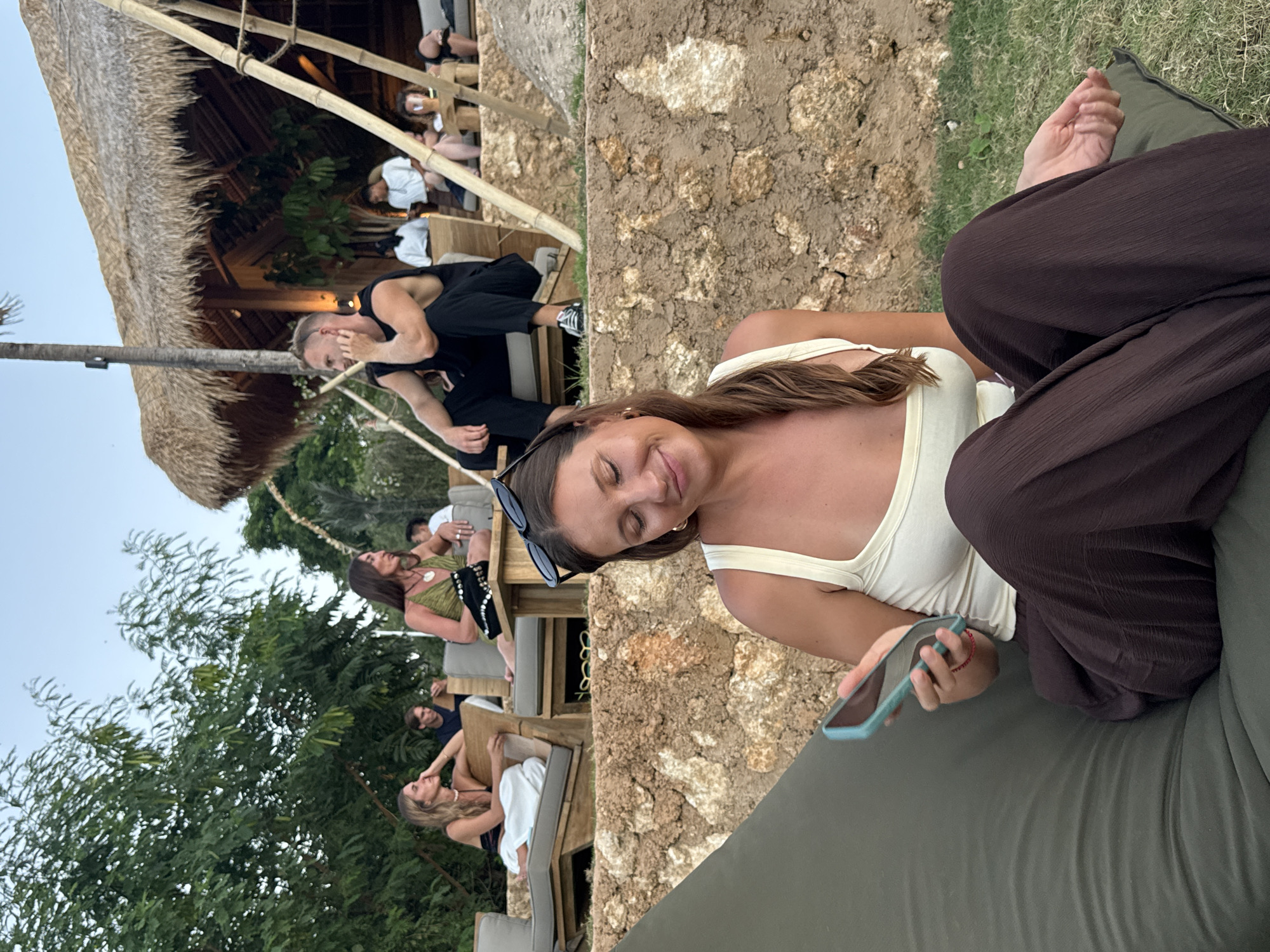 The image depicts a group of people gathered at an outdoor venue with a thatched roof structure and lush greenery. The scene appears to be a social gathering, possibly at a resort or restaurant, given the relaxed atmosphere and seating arrangements.
In the foreground, a woman is seated and looking at her phone. She is wearing a cream-colored top and dark brown pants. To her left, another person's hand and arm are visible, also appearing to be seated.
In the background, several other individuals are seen, some seated on benches and chairs, engaged in conversation or relaxation. The thatched roof and bamboo elements suggest a tropical or beachside setting, consistent with the provided location context of Kuta Selatan, Indonesia. The lighting indicates it is likely late afternoon or early evening. The overall mood seems casual and unhurried, with people enjoying each other's company in a scenic outdoor environment. There is no visible text in the image.