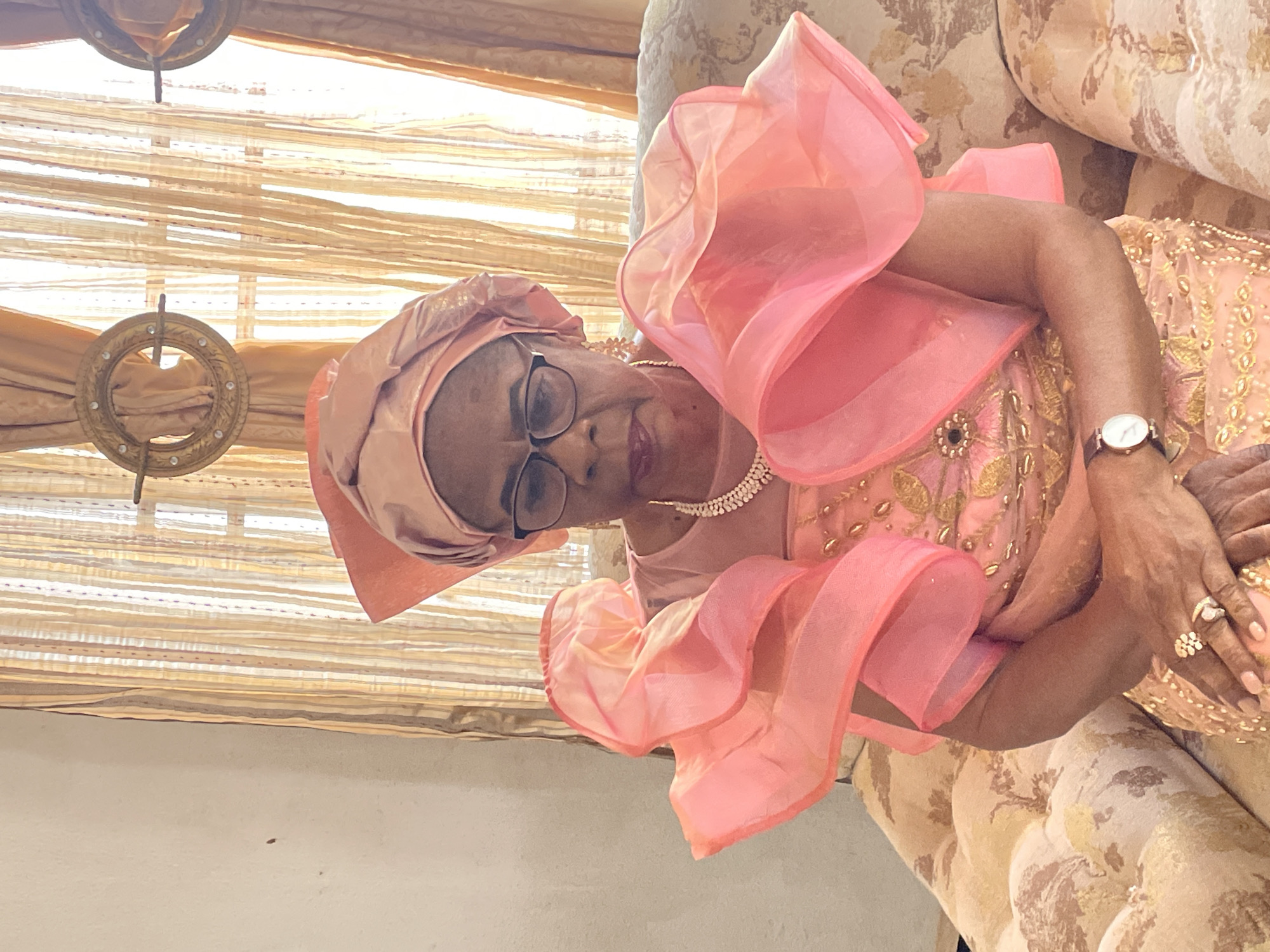 A woman wearing a pink dress and head wrap is seated on a patterned sofa. She is also wearing glasses, a necklace, a watch, and rings. The background includes a window with blinds and a decorative light fixture. The location is in the city of Unknown, Unknown.