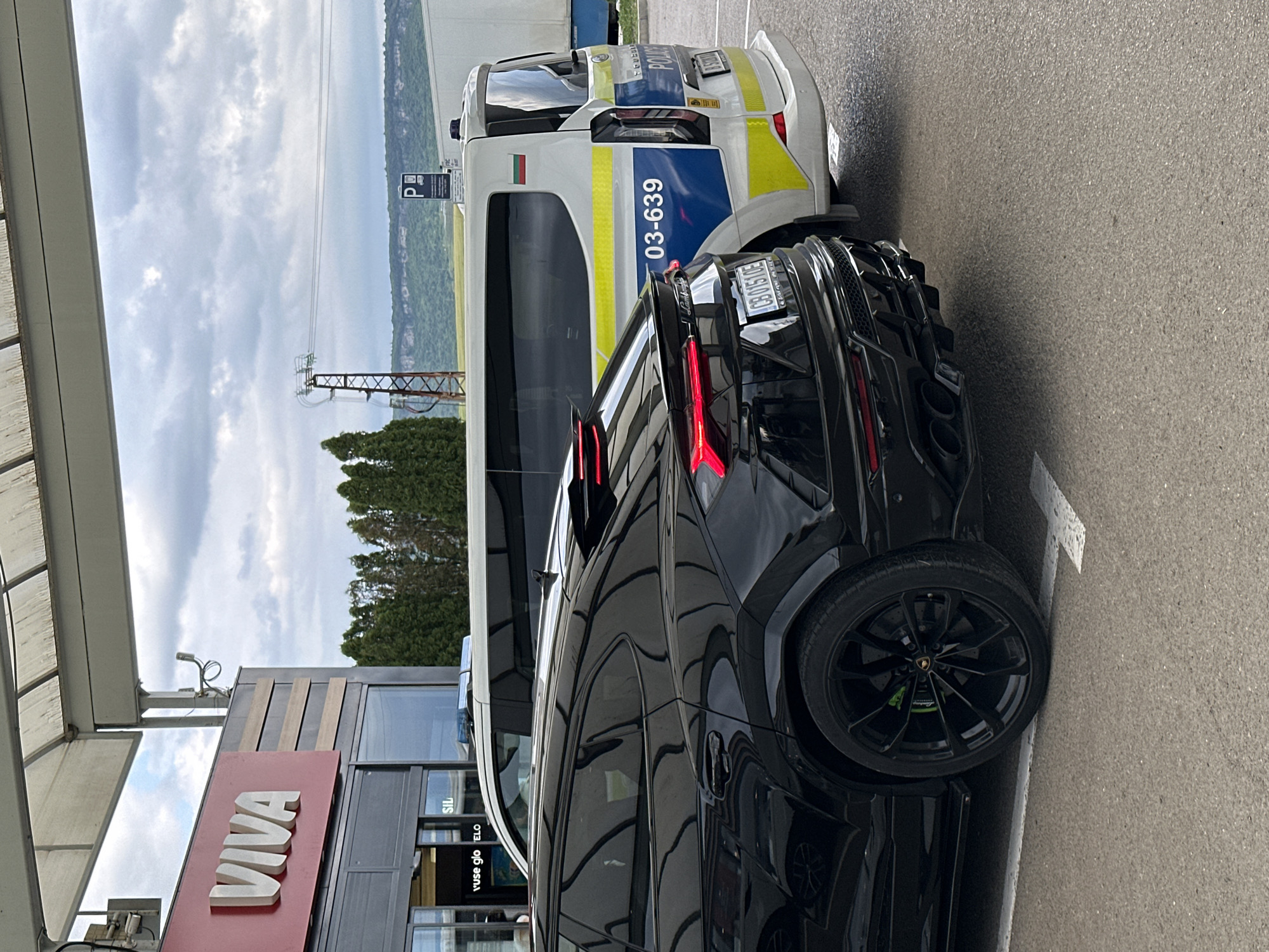 A black Lamborghini Urus and a Bulgarian police vehicle are parked next to each other at a VIVA gas station. The police vehicle is white with blue and yellow stripes and the number "03-639" displayed on its side. The Lamborghini has a license plate "CB 0150 UE". The surrounding environment includes a cloudy sky, trees, an electrical tower, and a hill in the background. The location is likely Kaspichan, Bulgaria, as the police car is Bulgarian.