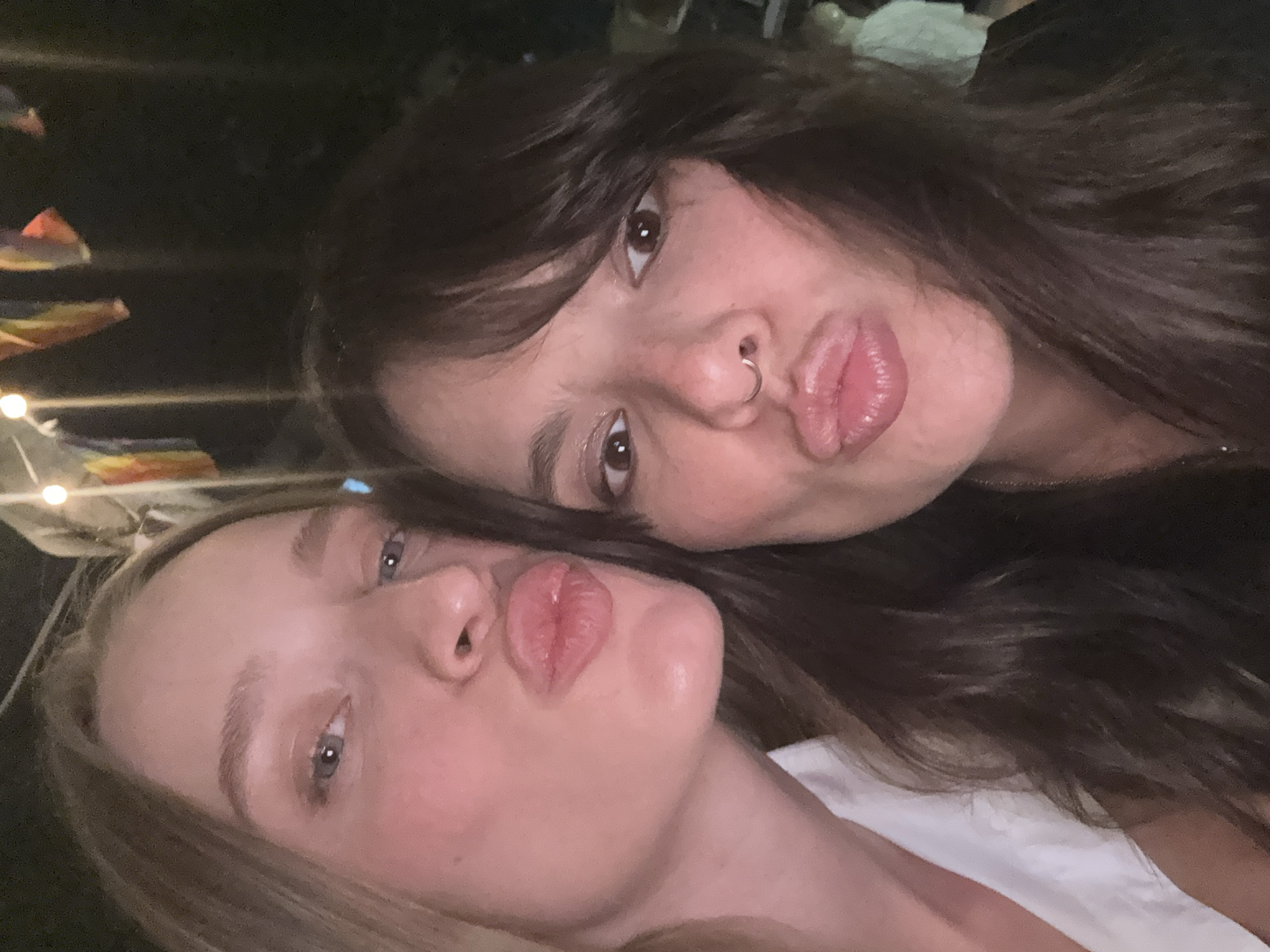 Two women are taking a selfie outdoors at night. The woman on the left has long blonde hair and is wearing a white shirt. She is puckering her lips. The woman on the right has dark hair and is wearing a black shirt. She also has her lips puckered. The background is dark with some out-of-focus lights and colorful flags hanging above. It is not possible to determine the specific location in Tel Aviv from the image.