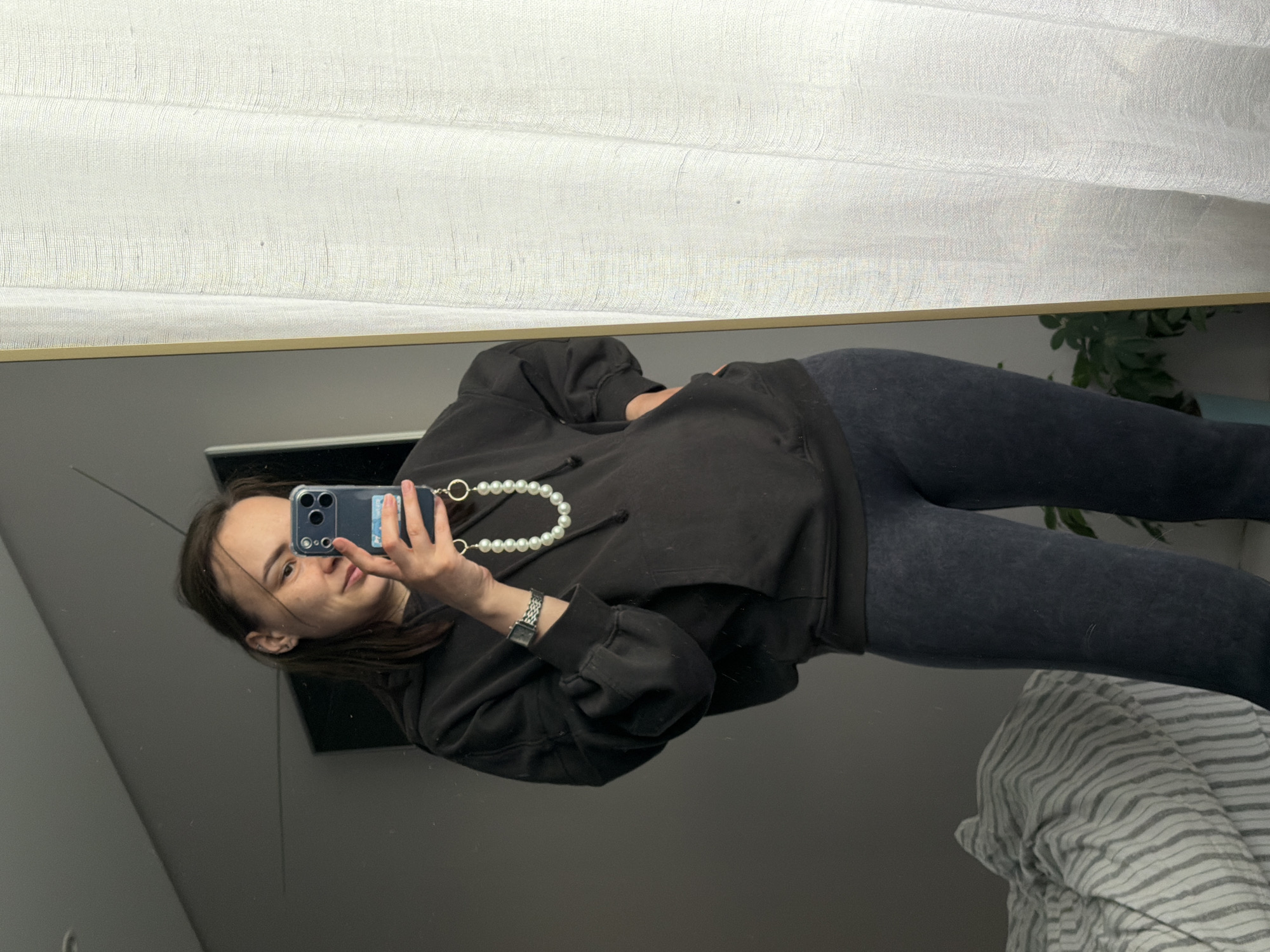 The image displays a mirror selfie of a young woman taken in an indoor setting, specified as Chaikovsky, Russia. The woman, with dark hair, is reflected in what appears to be a full-length mirror, holding a smartphone with a triple-lens camera array (resembling an iPhone) to capture the image. A strap made of white, pearl-like beads is attached to her phone case. She is dressed in a casual outfit consisting of a black, loose-fitting hoodie and dark, possibly charcoal-grey, leggings. A watch with a square face and a metallic link strap is visible on her left wrist.
She has a neutral to slightly smiling expression, looking directly towards the phone's camera. Above the mirror's reflection, a white, textured fabric, likely a curtain or blind, is visible, suggesting a window and a source of natural light. To the right side of the reflection, a green plant and striped bedding, possibly from a bed, can be seen. The area behind the woman in the reflection is a plain, dark surface, likely a wall. The photo is oriented sideways. No legible text is visible on any objects within the frame.