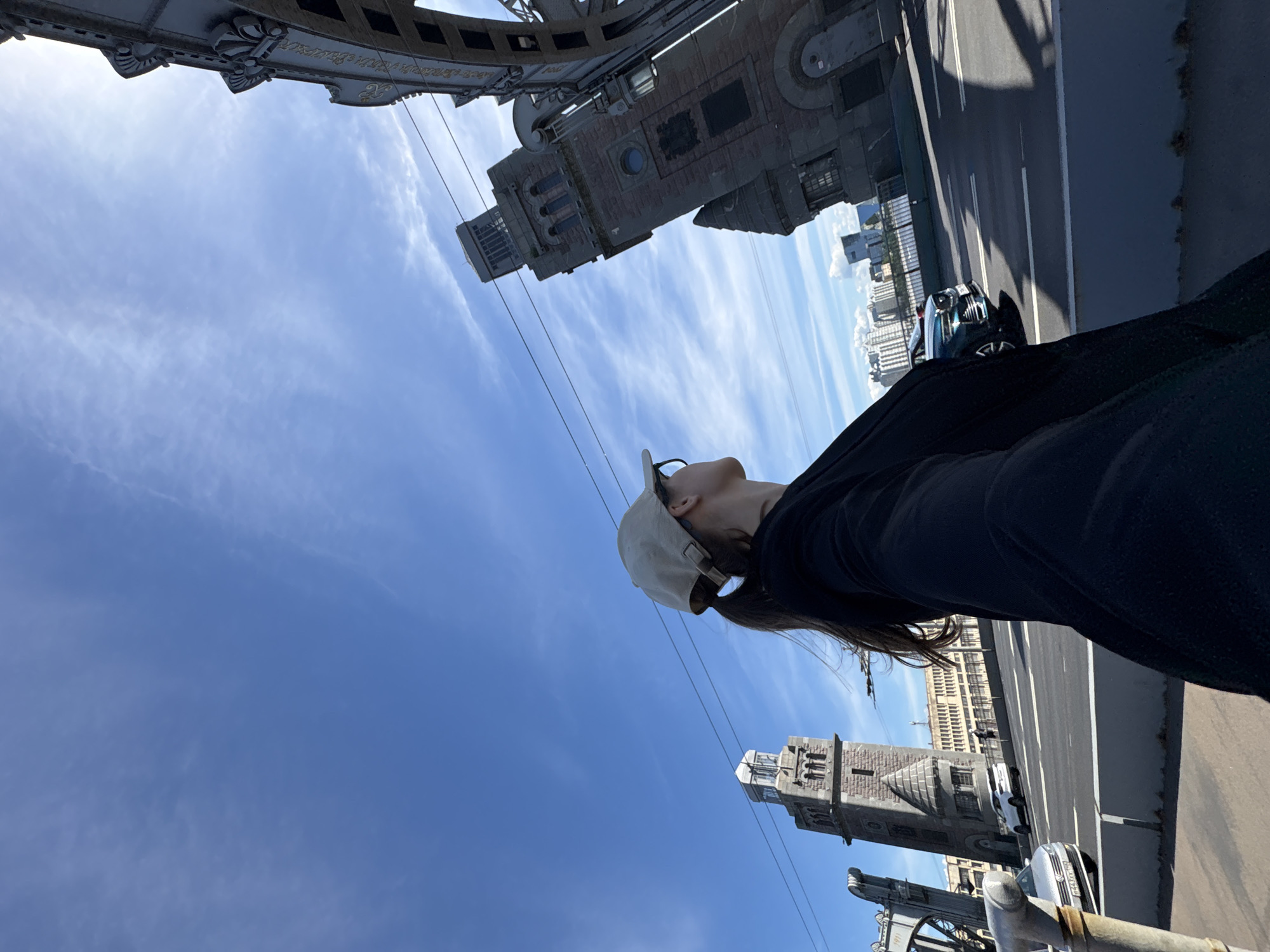 The media file is a photograph taken from a bridge in Saint Petersburg, Russia. The image is oriented such that the sky is predominantly visible, with a person in the foreground. The person is wearing a black top and a beige baseball cap, with their head tilted back and looking upwards. Their long, dark hair is visible cascading down.
The scene depicts a bright, sunny day with a clear blue sky and wispy white clouds. The perspective is from a high vantage point, looking down and across the bridge. Below the person, a road with traffic lanes is visible, with a few cars driving on it. Distinct architectural structures, characteristic of Saint Petersburg's bridges, are present in the background. These include ornate stone towers and intricate metalwork, suggesting a historic or grand bridge. One tower features a clock face. In the distance, buildings with multiple windows and a light-colored facade are visible, contributing to the urban landscape.
The overall mood conveyed is one of serene observation and perhaps contemplation, with the person seemingly enjoying the view and the weather. The sunlight indicates it is daytime, likely mid-morning or afternoon. There is no visible text in the image.