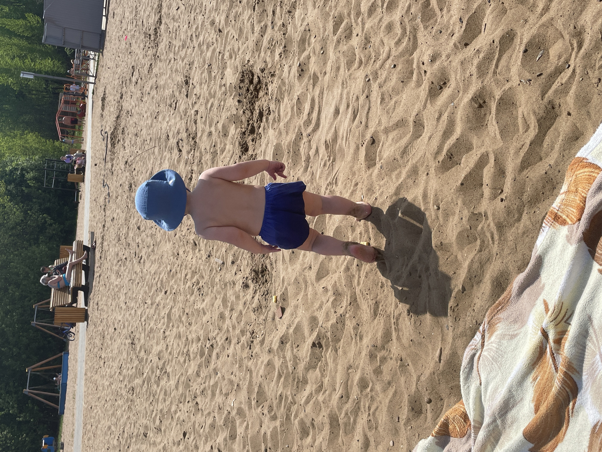 The image shows a child standing on a sandy beach. The child is wearing a blue hat and blue swim trunks. The beach appears to be located next to a park with benches and trees. The scene is brightly lit, suggesting it is daytime. The picture was taken in Perm, Russia. A tan and brown towel is folded on the right side of the frame.