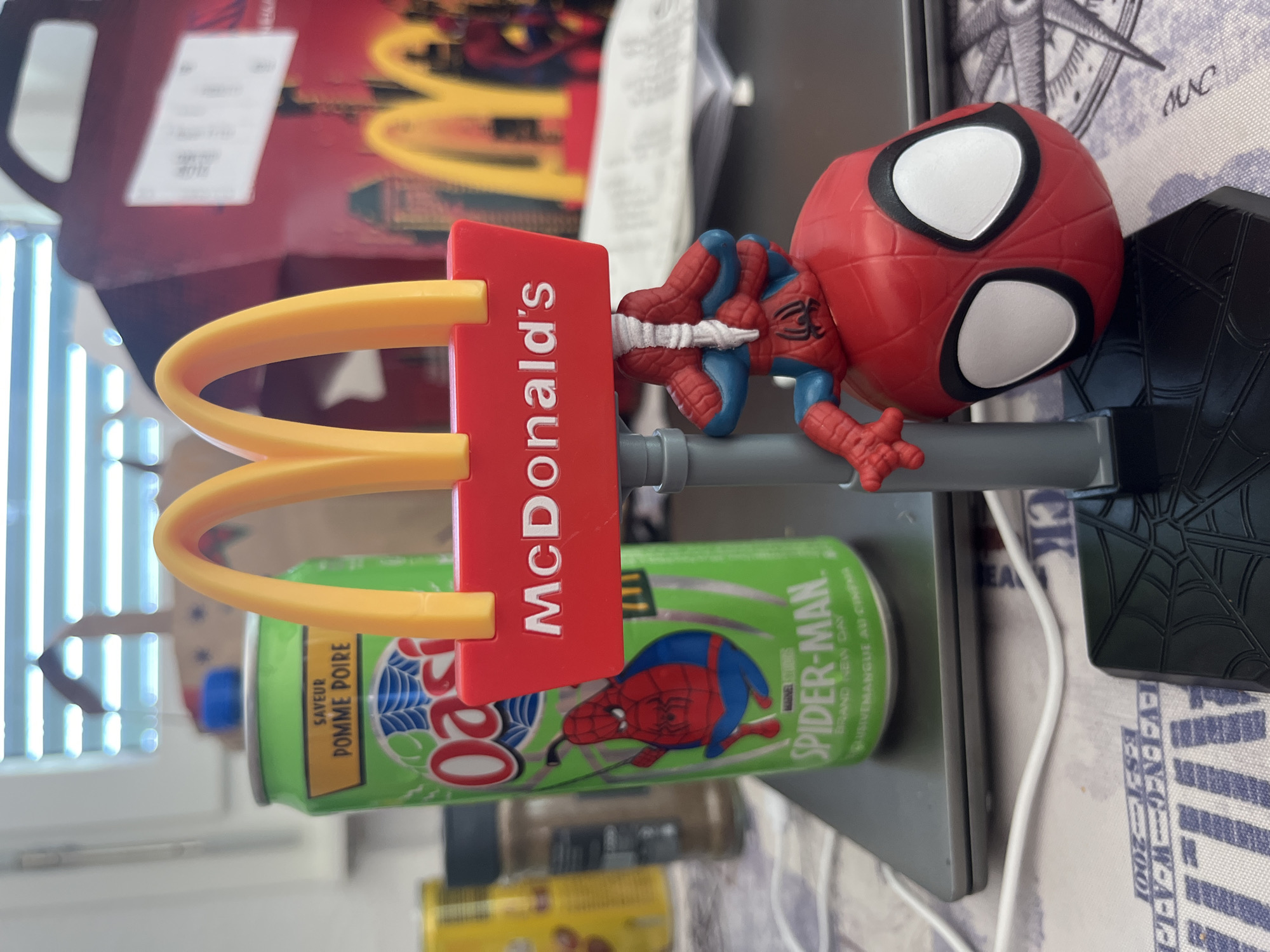 This image depicts a close-up of a McDonald's Happy Meal toy featuring Spider-Man, positioned in front of a can of Oasis apple-pear flavored drink. The McDonald's logo, with its iconic golden arches, is prominently displayed on a red sign with the text "McDonald's" in white lettering. The Spider-Man figure is hanging from the sign, with its head resting on a black base adorned with a spiderweb pattern. In the background, a blurred McDonald's Happy Meal box is visible, along with other out-of-focus objects on a table covered with a patterned cloth. The scene appears to be indoors, with natural light coming from a window with blinds in the upper left corner. There is no indication of the city of Geneva, Switzerland, or any specific location within it.