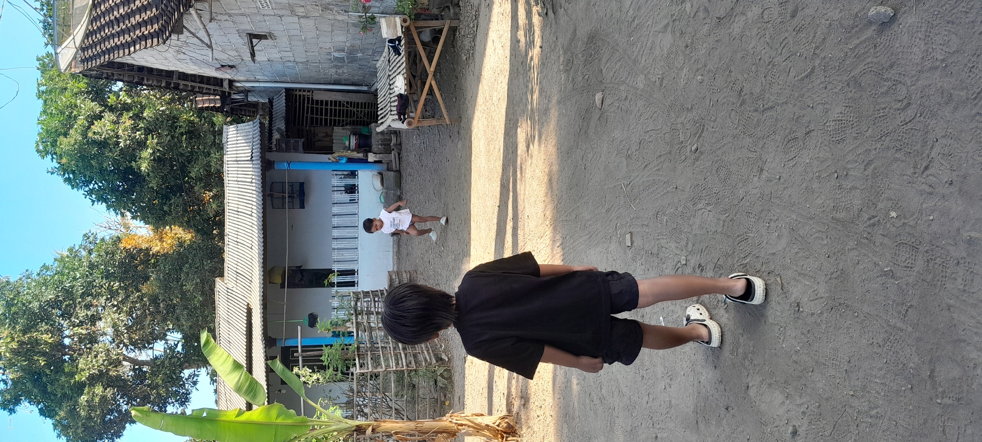 In Gintangan, Indonesia, two young boys are in a dusty yard in front of a modest house. The boy in the foreground is wearing a black t-shirt and black shorts, with white and black clogs. He is facing away from the camera, looking towards the other boy. The second boy, further in the yard, is wearing a white t-shirt and appears to be in motion. The house has a corrugated metal roof and a light-colored exterior. A bamboo bench with some items on it is to the right of the frame. Trees surround the area, and the sky is clear and blue. The ground is dirt with numerous visible footprints.